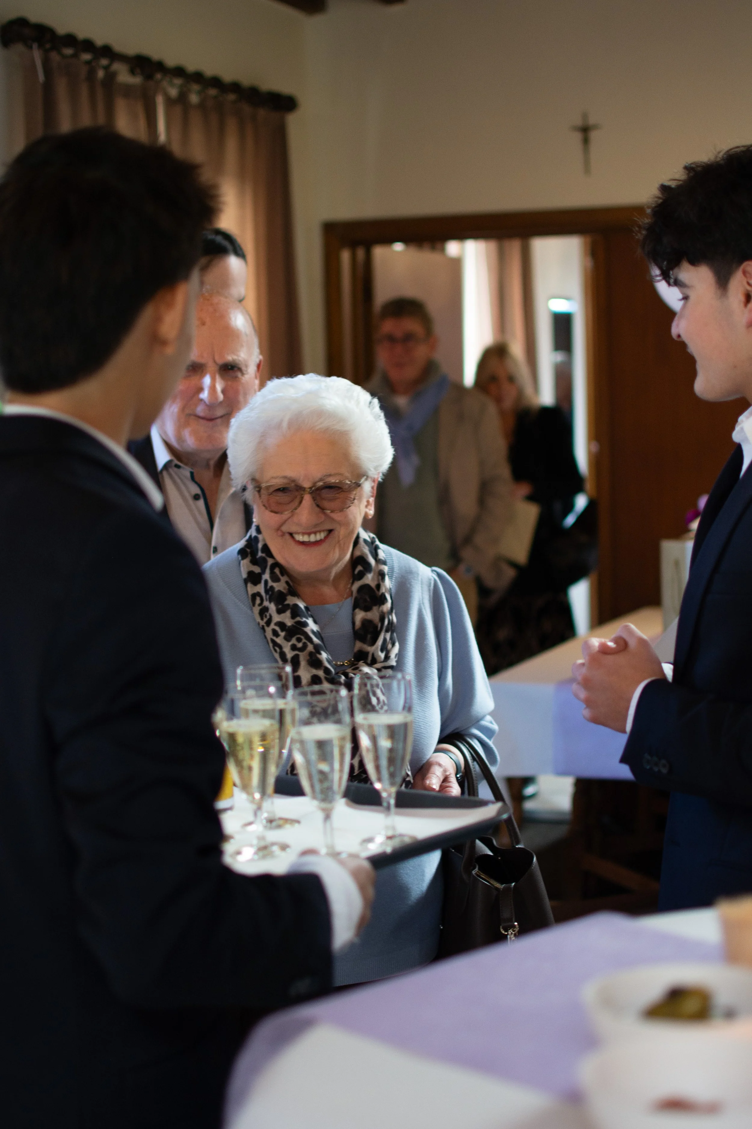 An elderly woman in glasses and a leopard print scarf smiling and talking with a young man at a social gathering, with others in the background and glasses of champagne on a tray.