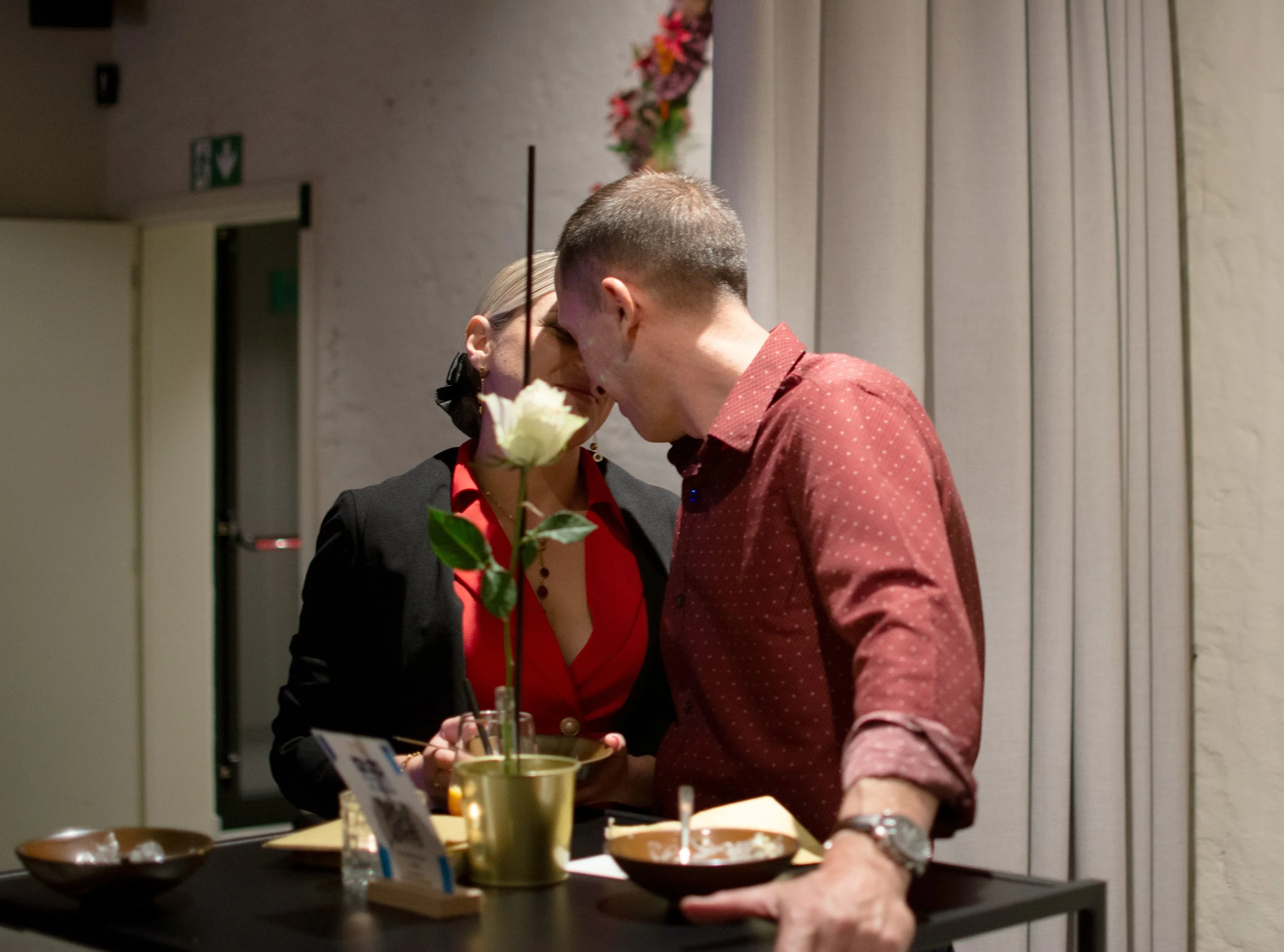 A man and woman are sharing a close moment, leaning their foreheads together, at a table indoors with a pink flower arrangement in the background.