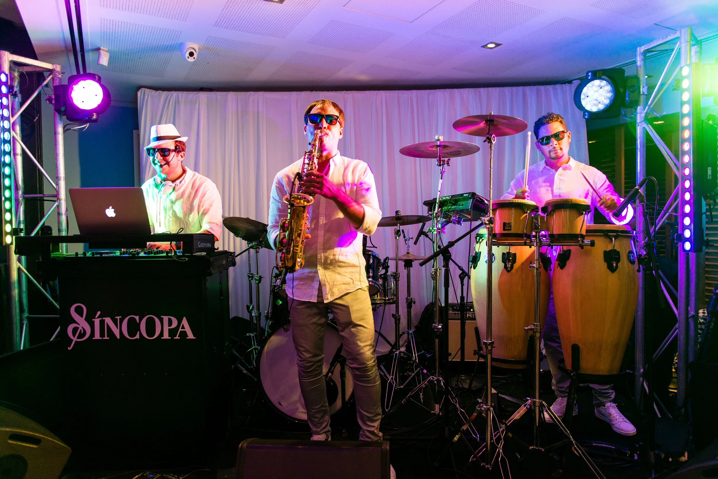 A band of three musicians performing on stage with colorful lighting. One is DJing with a laptop, another is playing saxophone, and the third is playing percussion on bongos. They are dressed in white shirts and sunglasses, with a backdrop of white curtains.