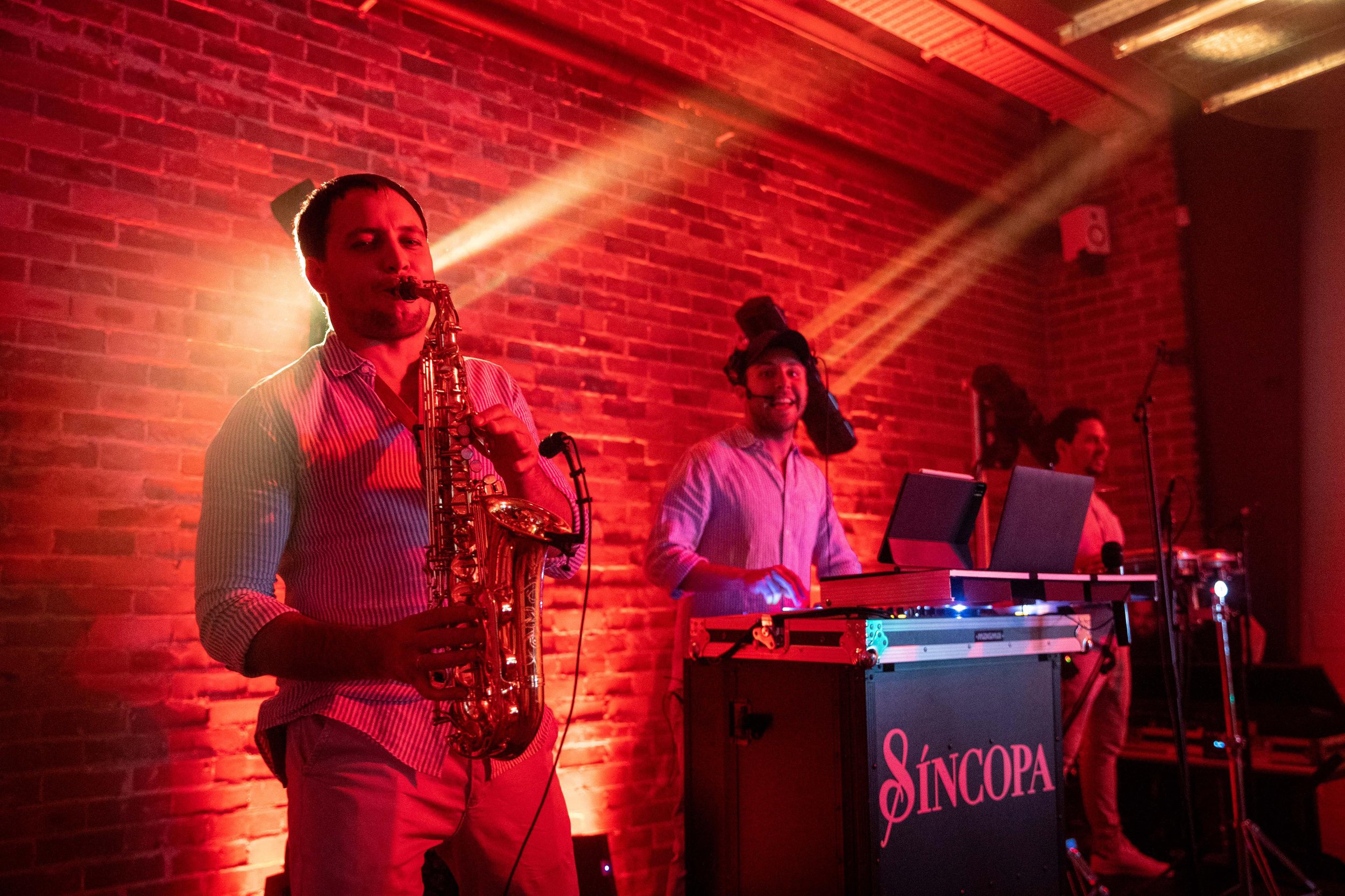 A band performing live on stage against a brick wall illuminated with red and orange lighting. The drummer is visible in the background, and two musicians, one playing a saxophone and the other at a laptop, are in the foreground.