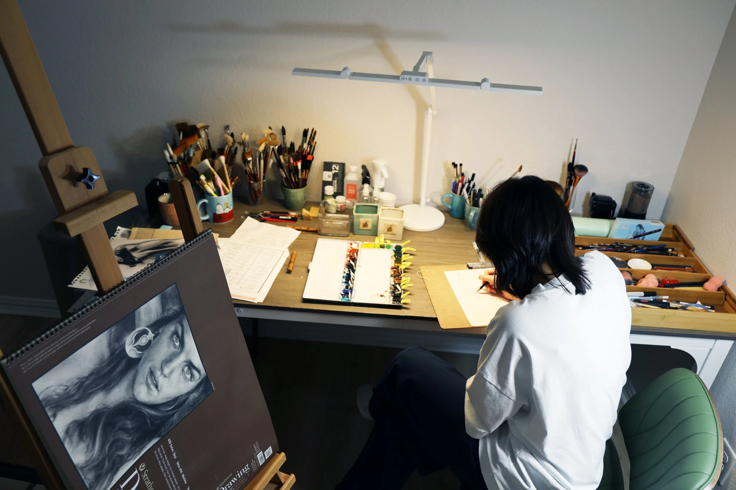 A person with shoulder-length black hair drawing at a cluttered art workspace with brushes, paints, sketches, and supplies on a wooden desk, with a portrait drawing of a woman on an easel.