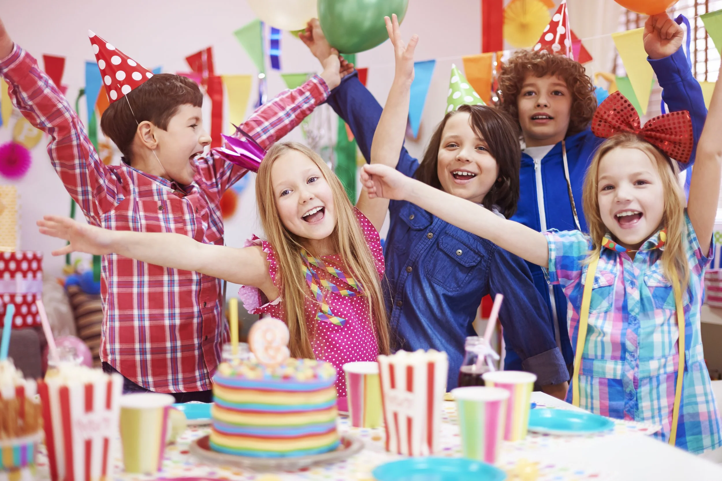Children celebrating a birthday with colorful party hats, balloons, and a decorated table with a birthday cake and snacks, smiling and joyfully raising their hands.