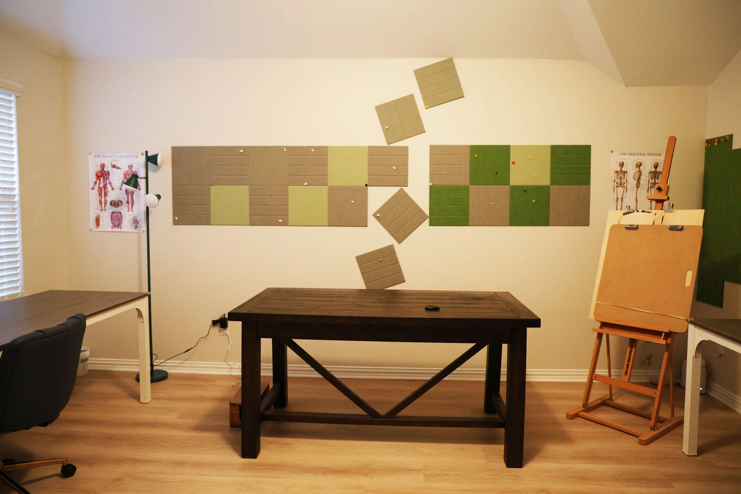 A room with beige walls, wooden floor, and various wall-mounted panels in lichen green, beige, and brown. There is a black chair on the left, a small wooden table in the center, and an easel with a sketchpad on the right. A poster on the far left dep