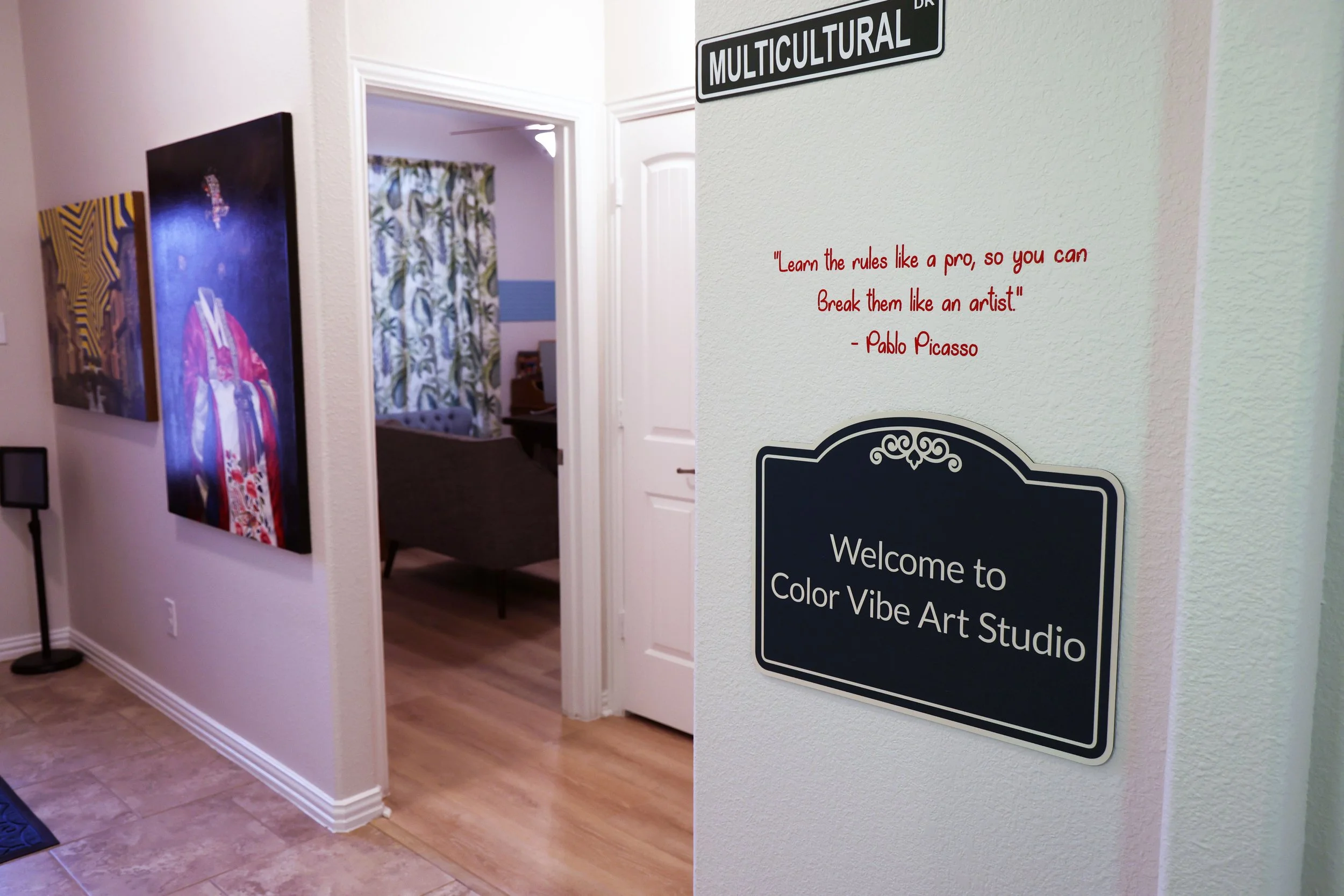 Interior of an art studio with paintings on the wall, a sign saying "Welcome to Color Vibe Art Studio," a quote by Pablo Picasso in red text, and a Multicultural Drive street sign.