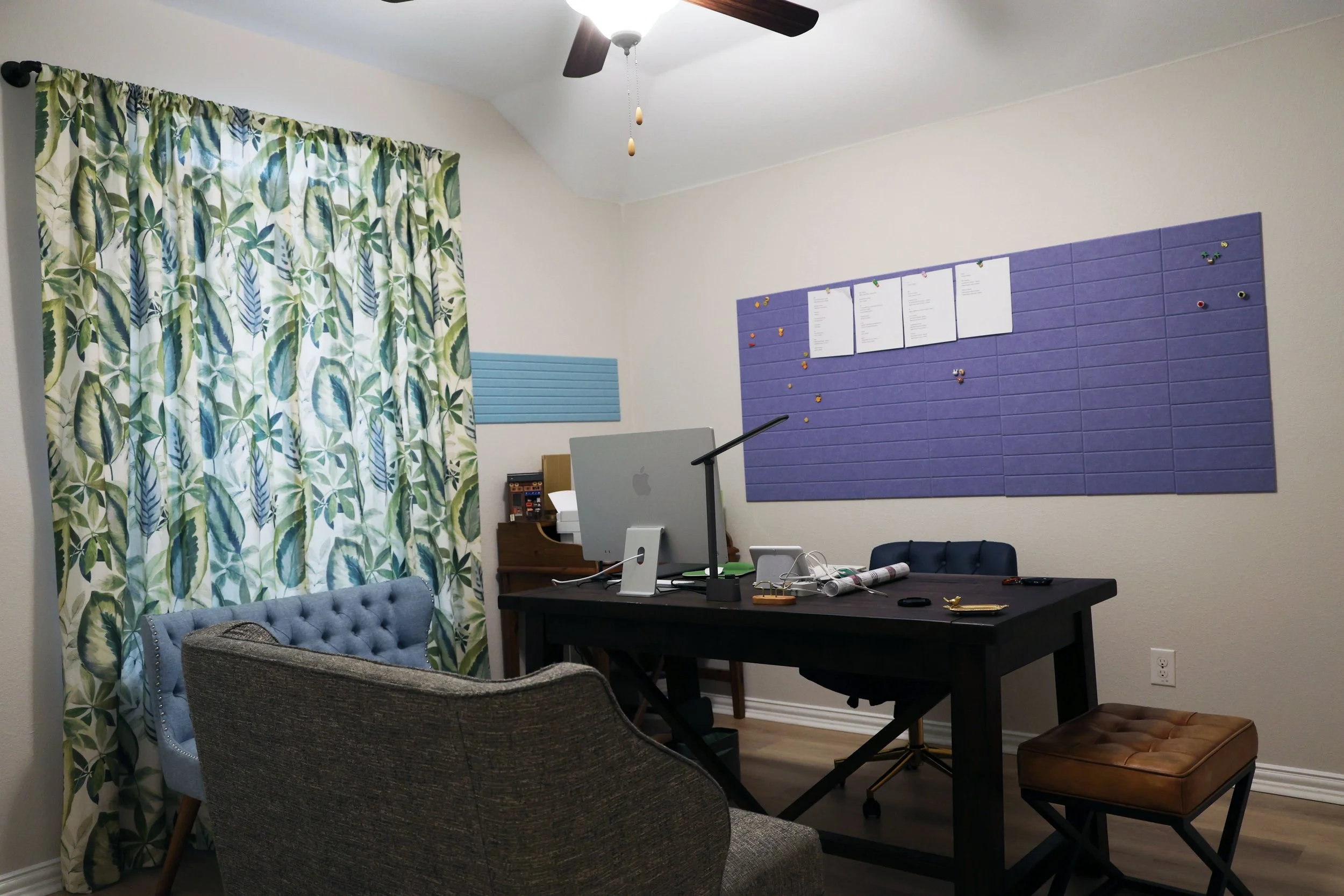 Home office with a black desk, blue tufted armchair, gray upholstered chair, brown leather ottoman, potted plant, patterned curtains, and a purple bulletin board on the wall.
