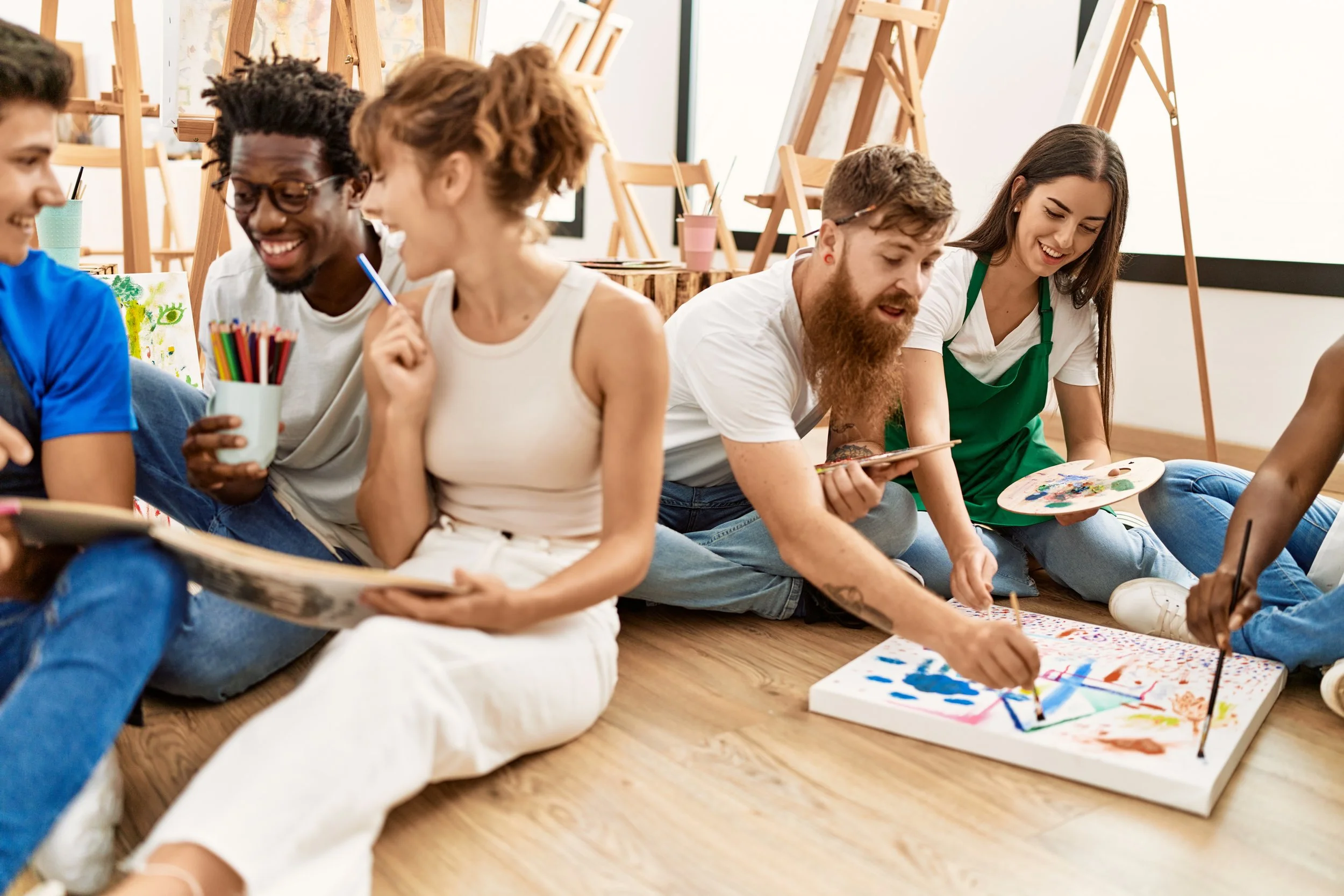 Group of young adults sitting on the floor of an art studio, engaging in painting and art activities, with easels and art supplies visible in the background.