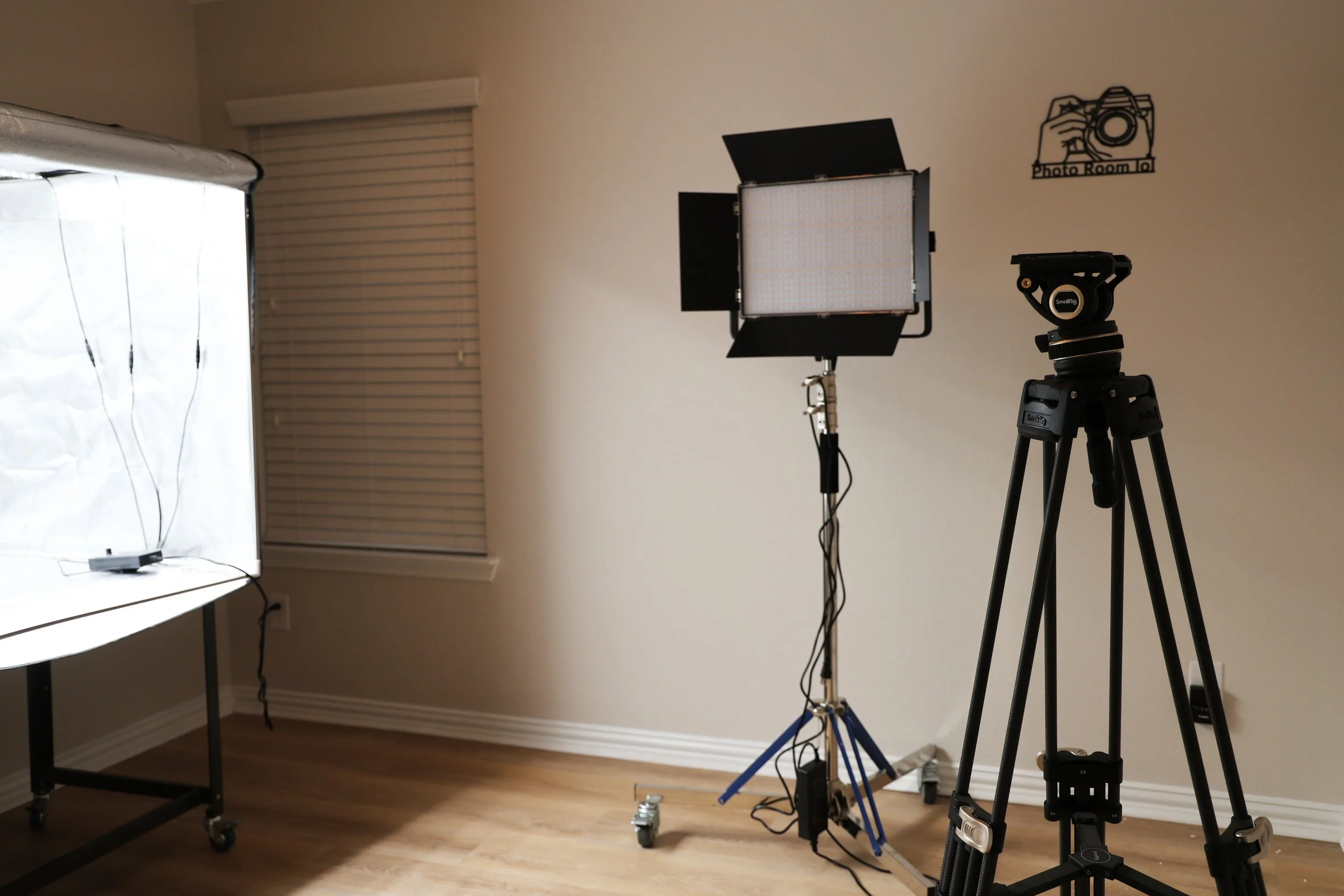 Photography studio setup with a large softbox light, a camera on a tripod, and a backdrop on a table with lighting cords, in a room with a window and blinds.