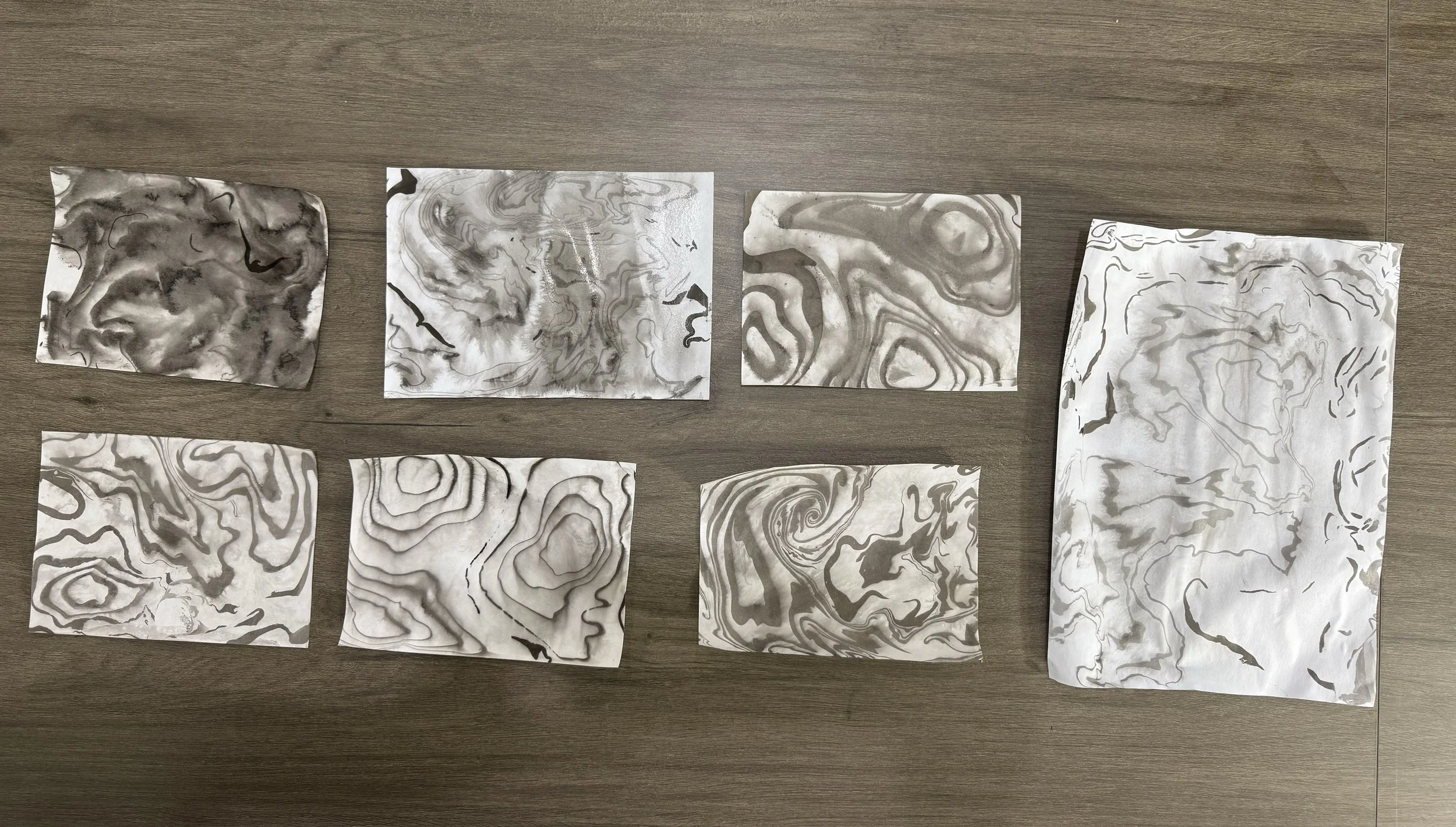 Eight sheets of paper with black and gray marbled patterns are arranged on a wooden surface. The sheets vary in size and pattern, with some being rectangular and one larger sheet on the right.