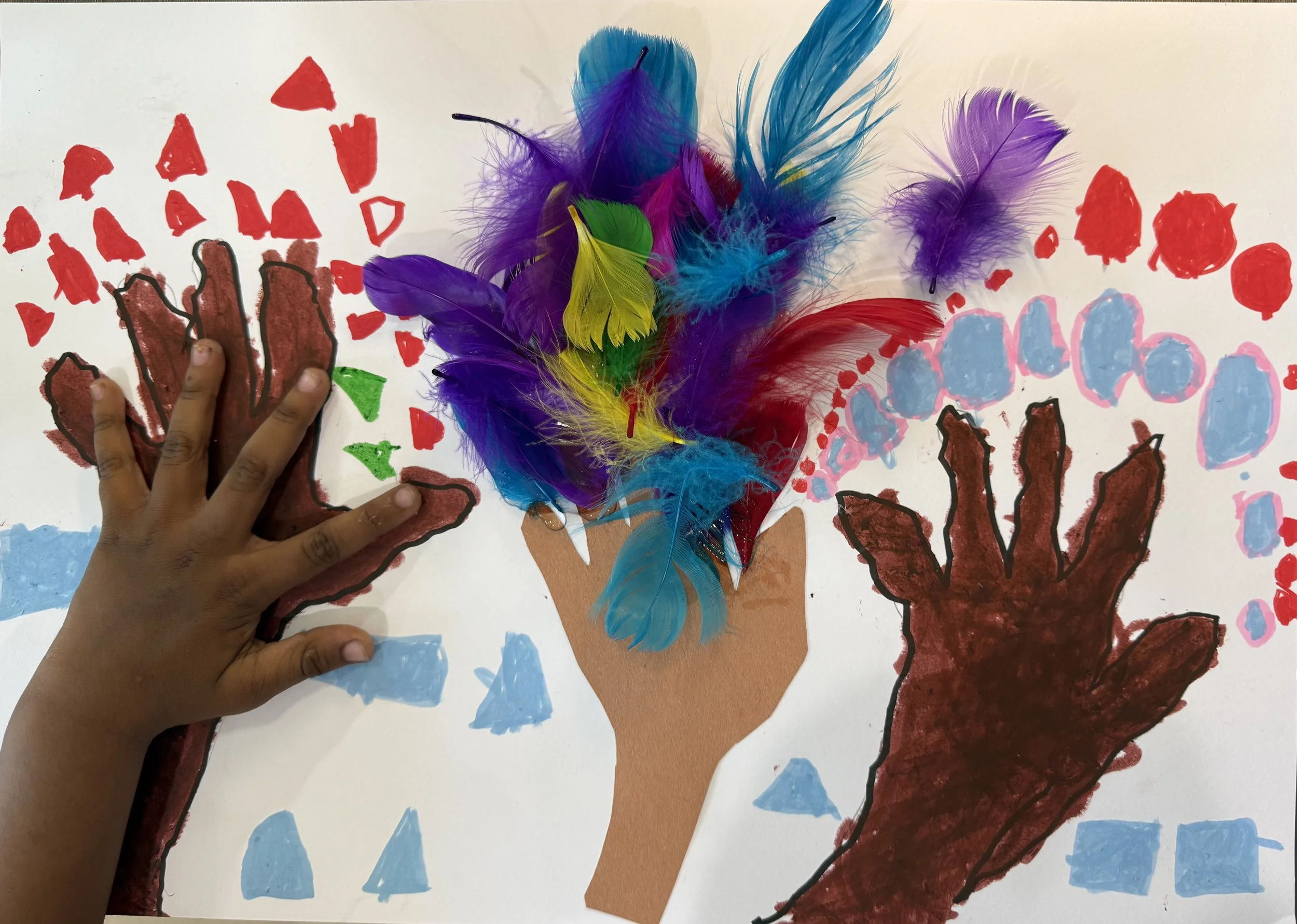 A child's hand with four fingers is resting on a colorful art project on paper. The artwork includes orange, red, green, blue, purple, and yellow paint and feathers, with handprint outlines and various shapes, set against a white background.