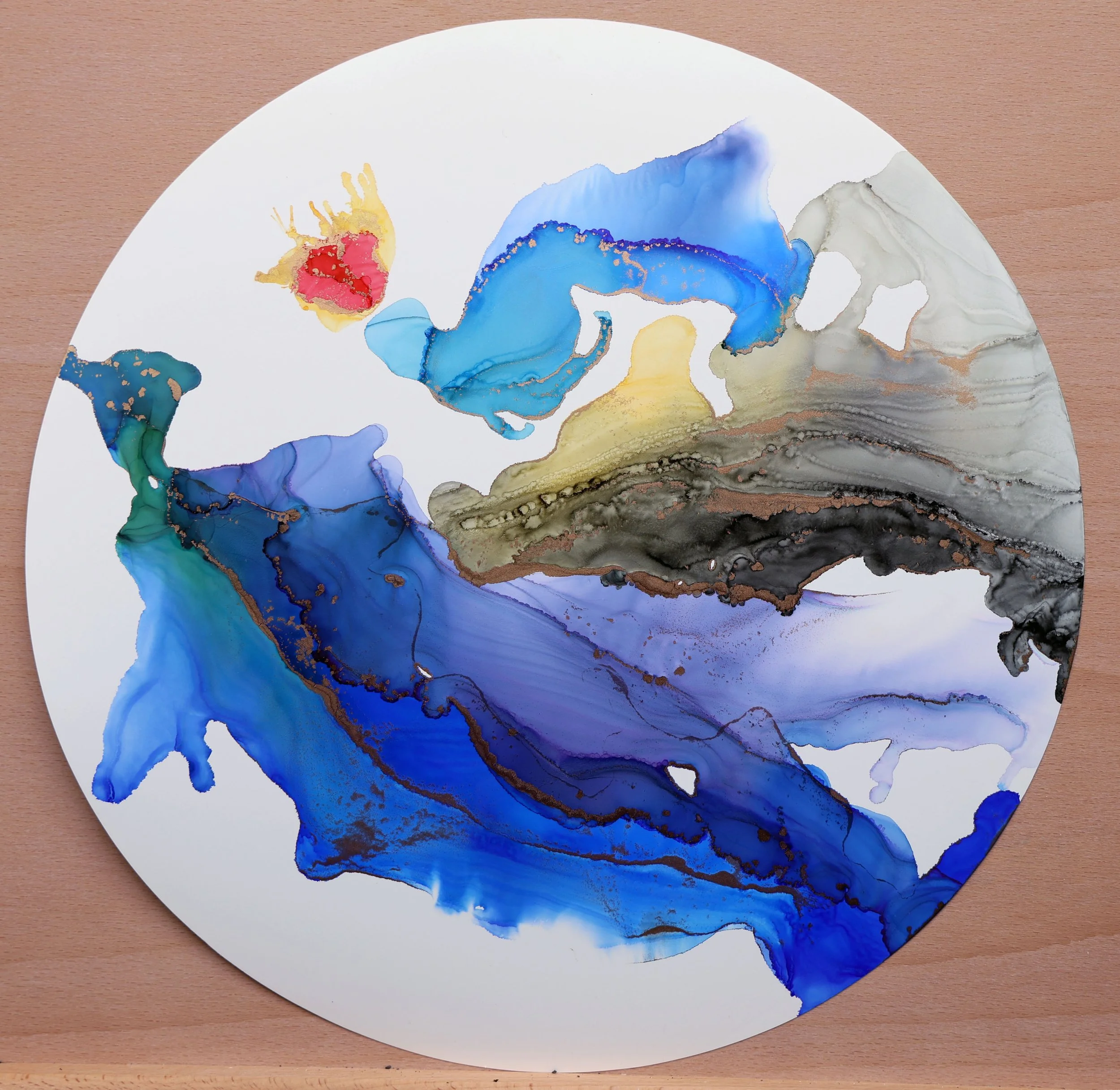 Abstract watercolor painting with blue, black, gray, yellow, red, green, and purple colors on a circular white paper, placed on a wooden surface.