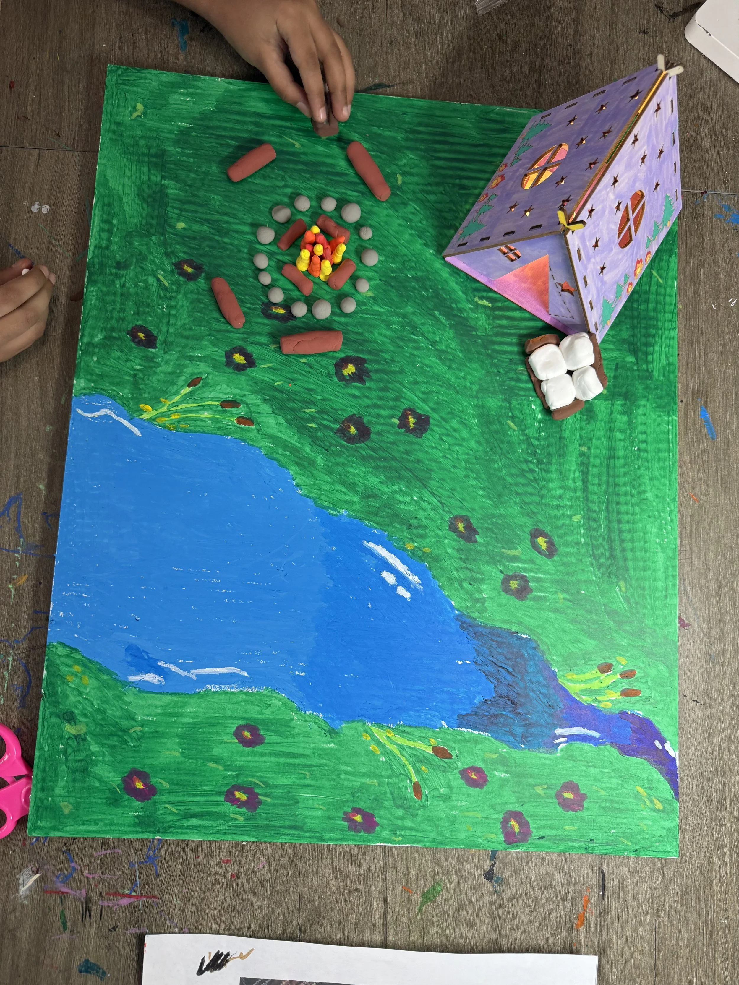A painted landscape scene on a large piece of paper with a blue river, green grass and purple flowers, a purple decorated house with cut-out windows and a brown door, and two small white houses on a grassy patch. Red and yellow toy pieces are placed 