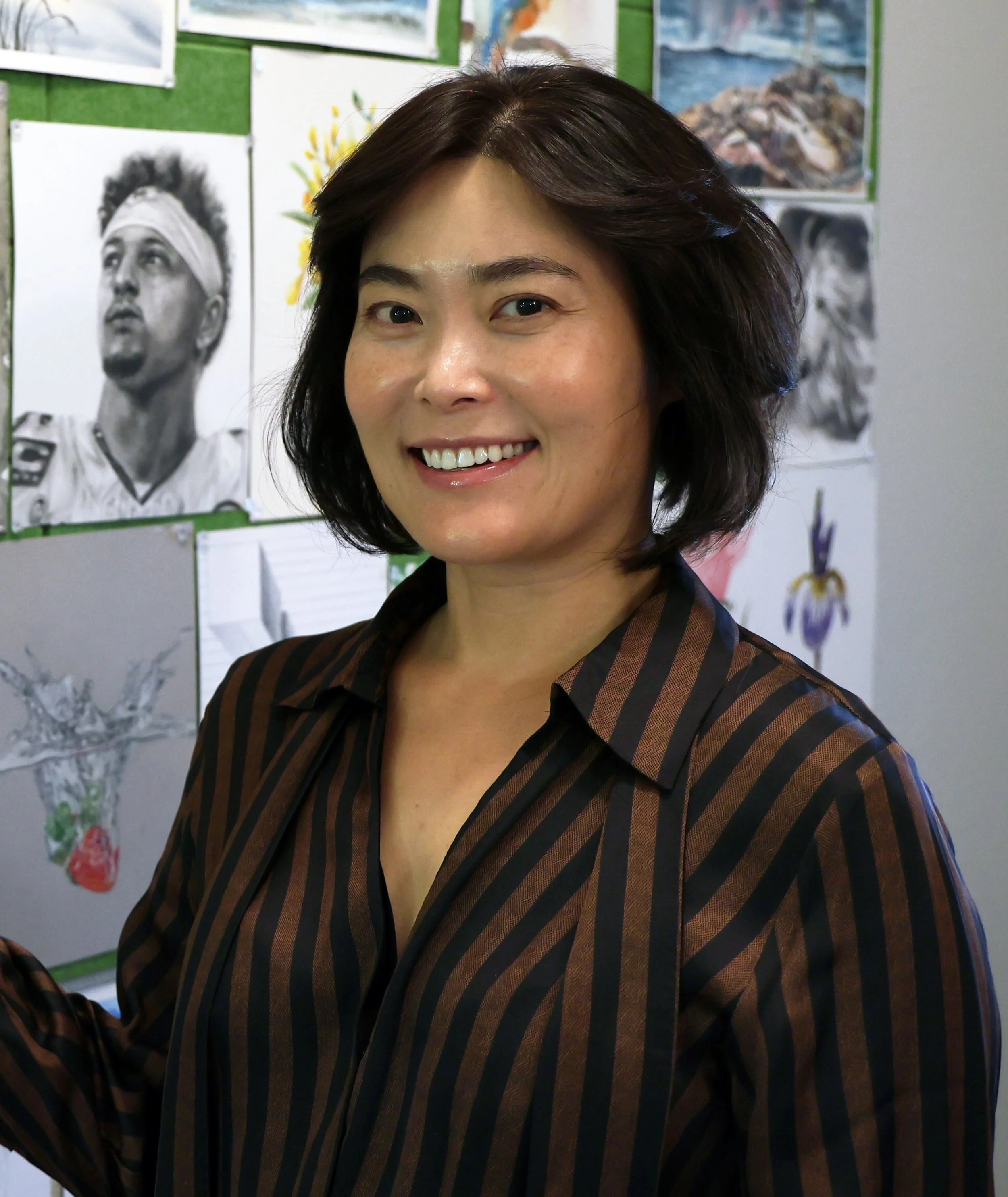 A woman with short dark hair smiling, standing in front of a bulletin board with various photos and drawings.