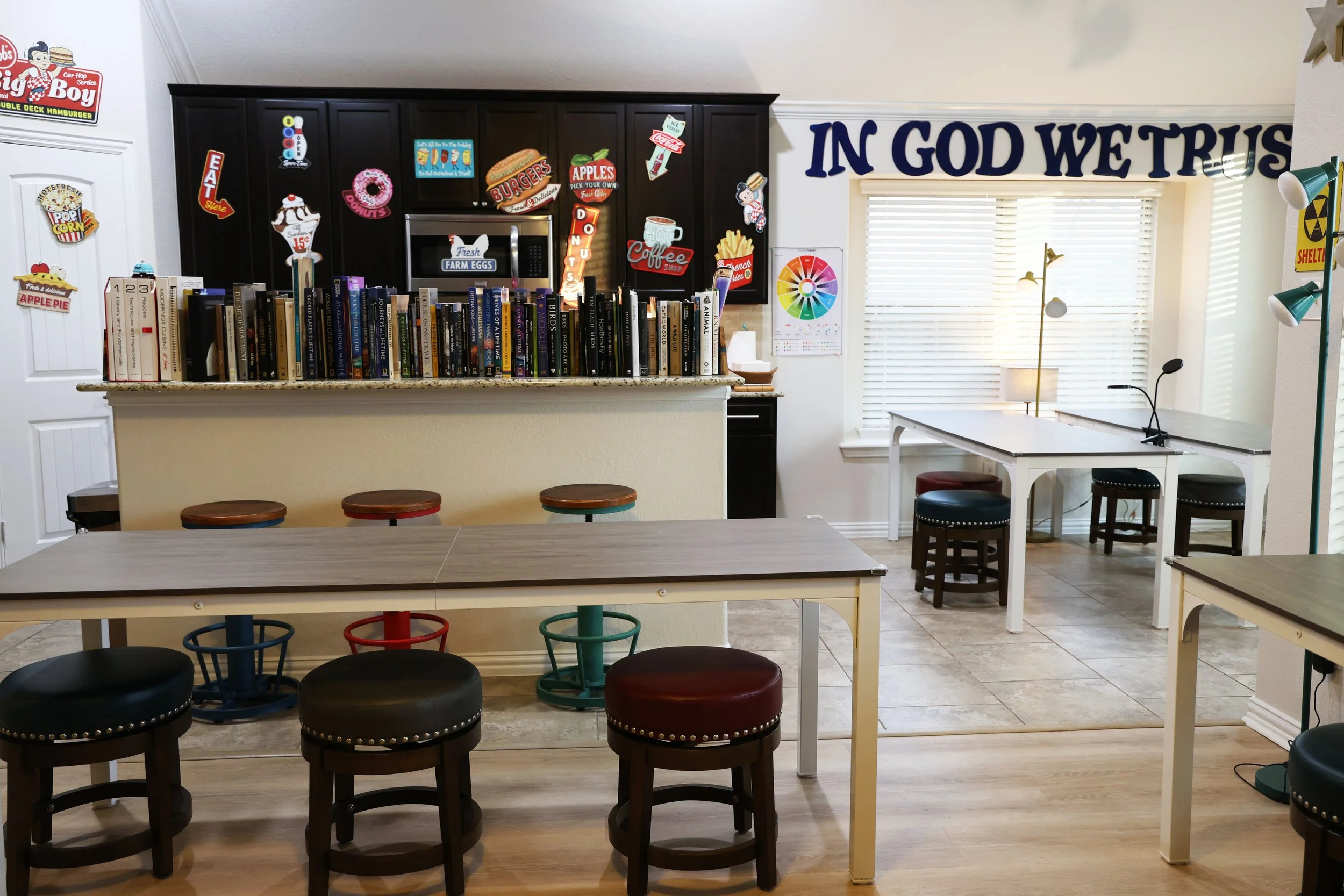 A cozy cafe or library with tables and chairs, a bookshelf with books, and decorative signs and posters on the walls, including a large sign that says 'IN GOD WE TRUST'.