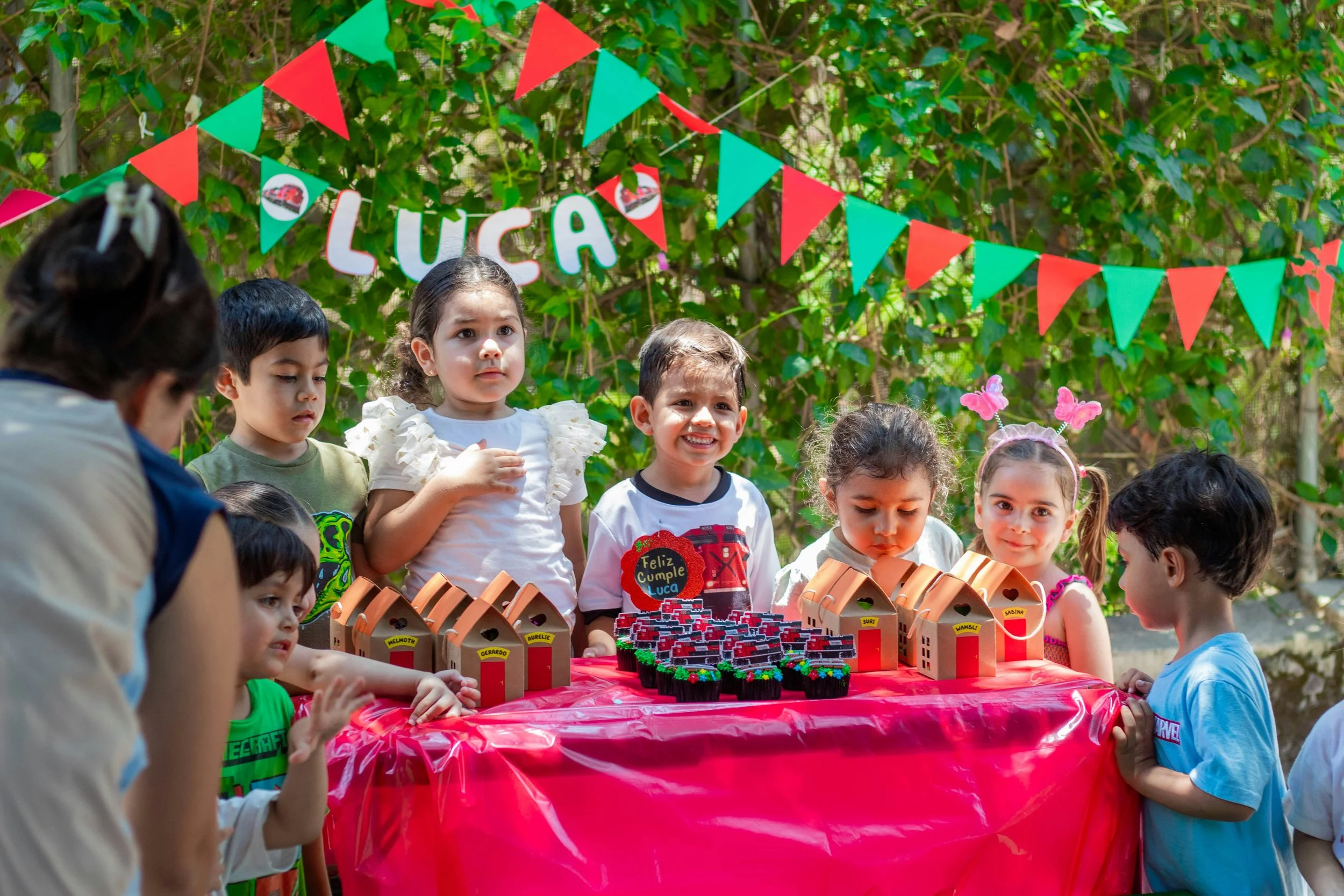 Children gathered around a birthday table outdoors, celebrating Luca's birthday with cupcakes and gift boxes, decorated with red and green bunting and a sign that says 'Luca'.