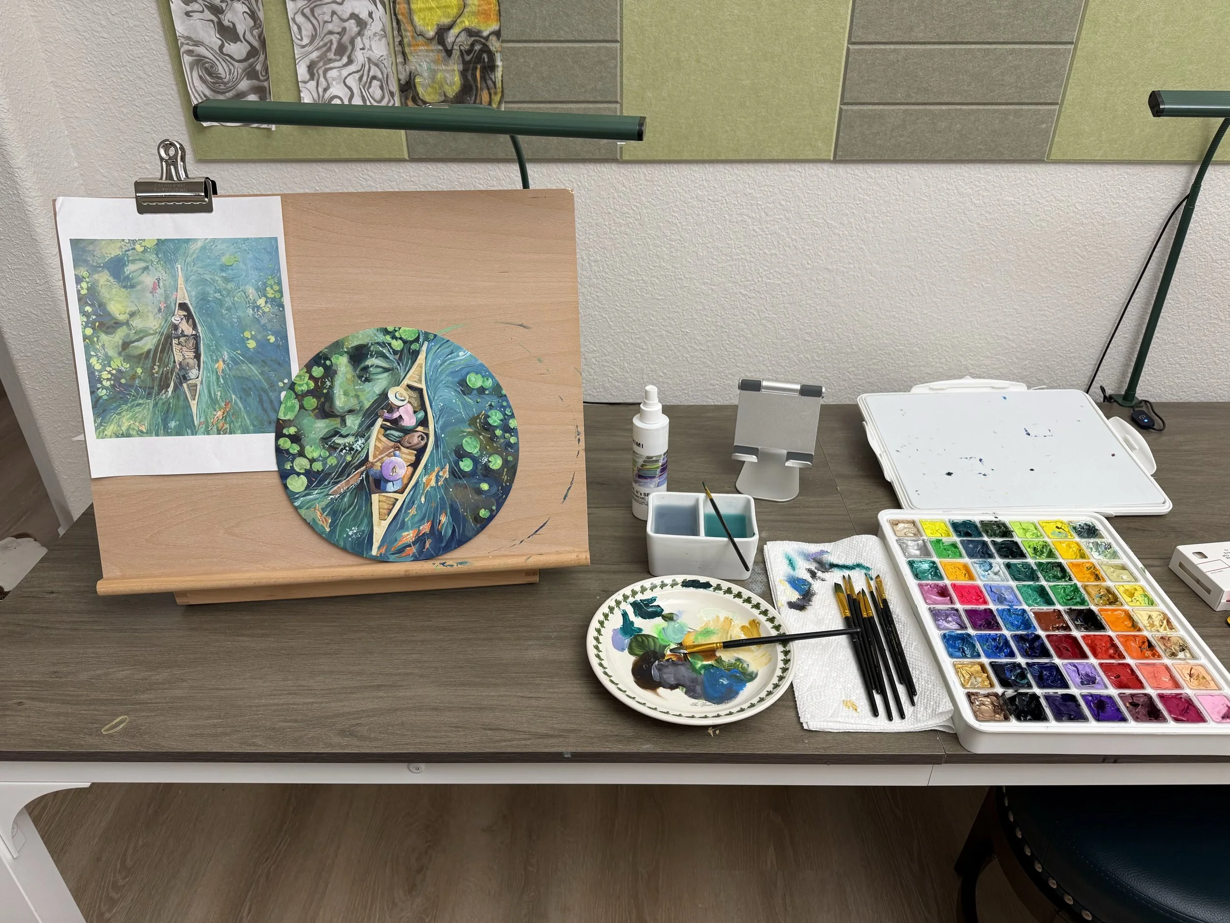 An art workspace with a painting setup. A completed watercolor portrait and a smaller version of the same painting on a wooden board. Art supplies include paintbrushes, a palette with mixed colors, a tray of watercolor paints, small containers with w
