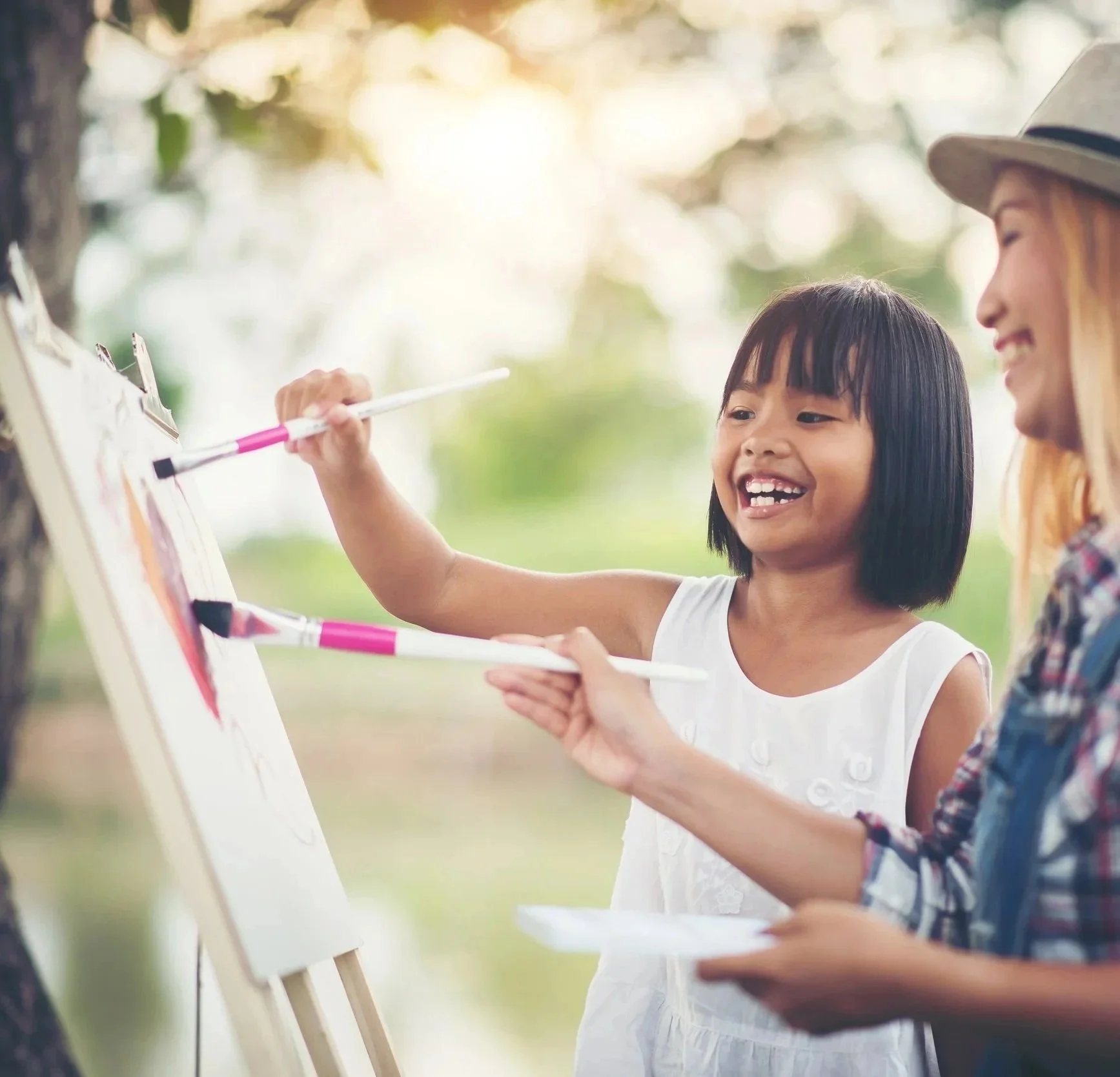 A young girl smiling while painting outdoors with an adult woman, both holding paintbrushes and working on a whiteboard.