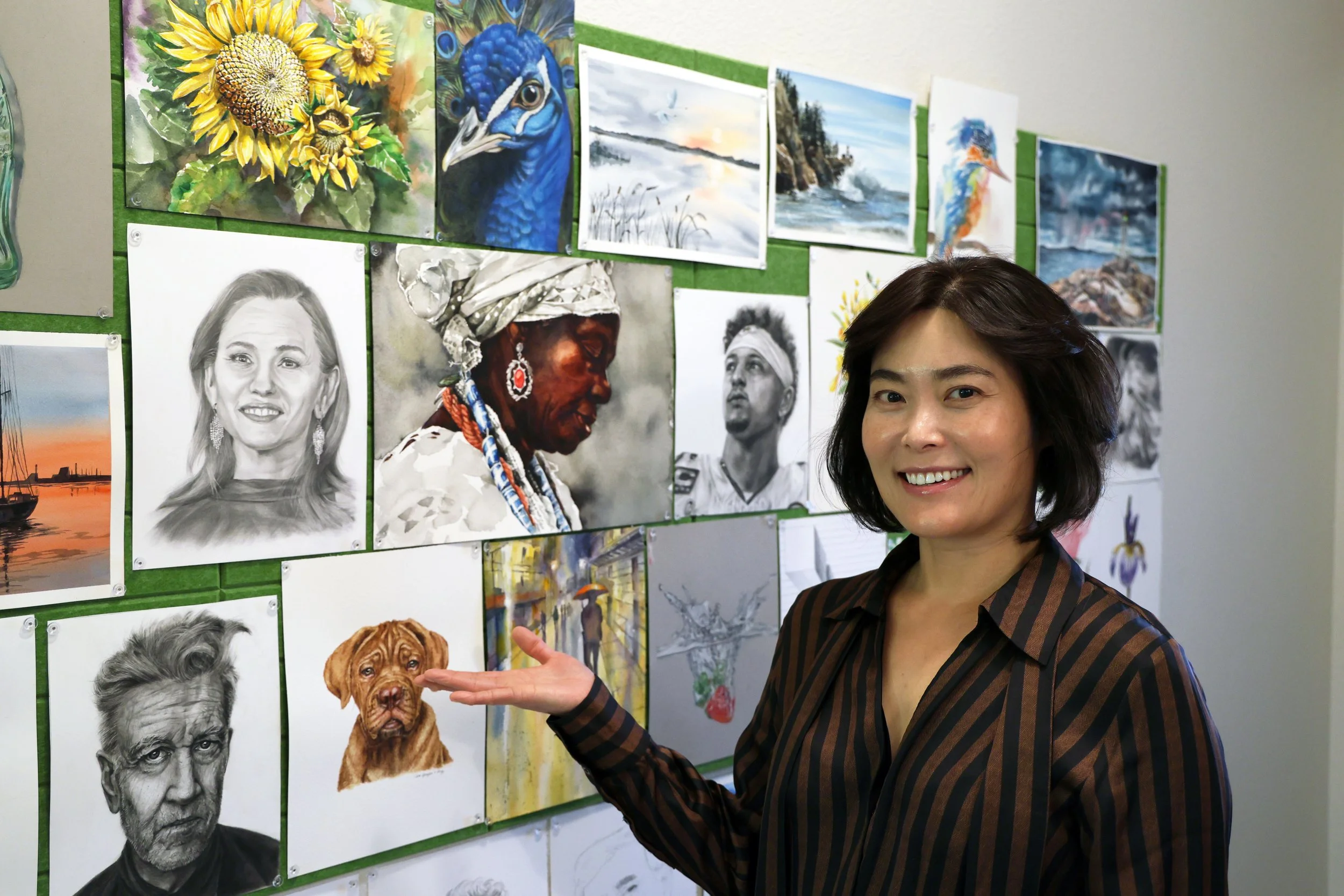 A woman stands in front of a wall with various artworks, including pencil sketches and colorful paintings of animals, landscapes, and people, pointing at a drawing of a brown puppy.