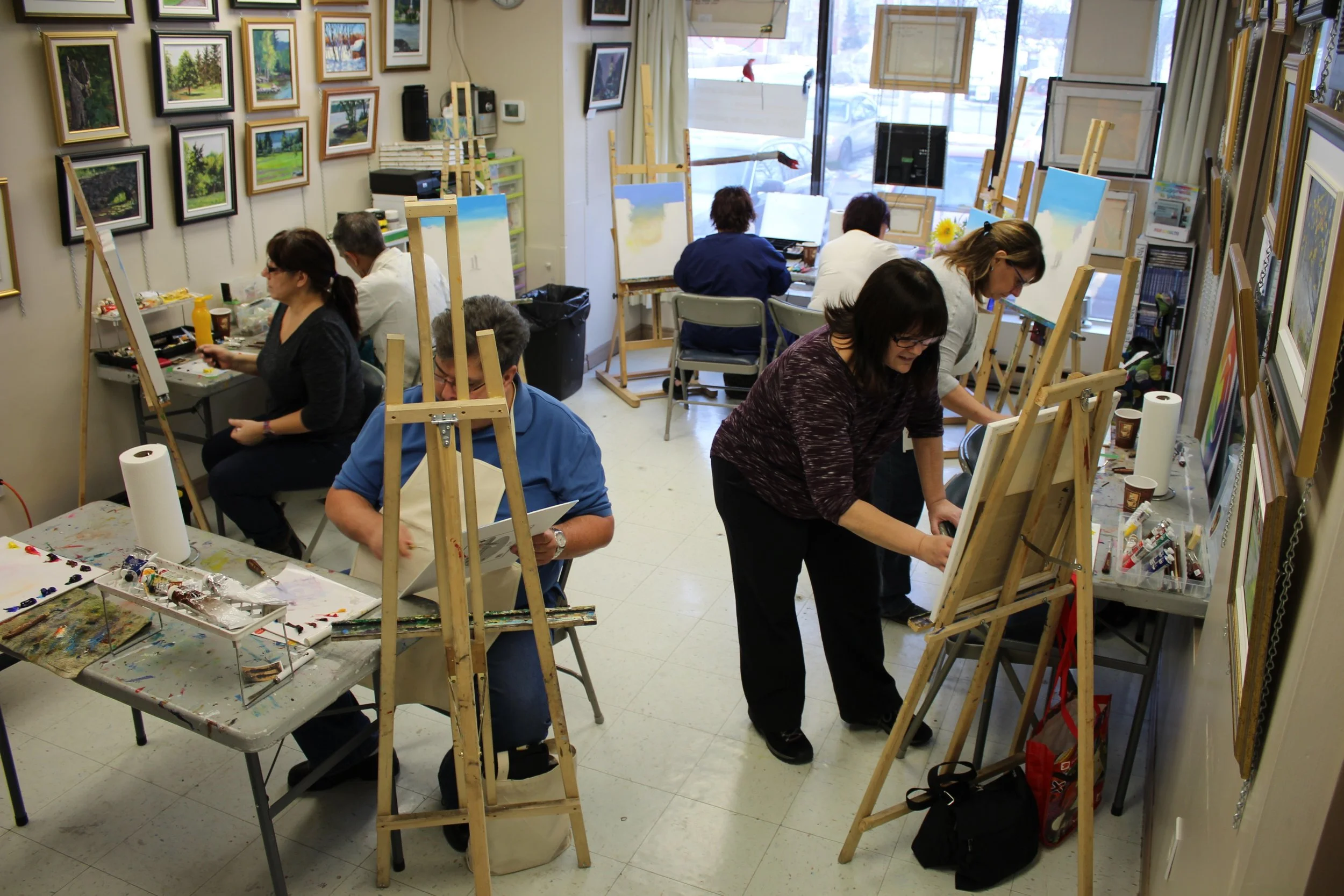 A group of people painting on canvases in an art classroom. Several easels are set up, and some people are working at tables with art supplies. Framed landscape paintings are on the walls, and a large window provides natural light.