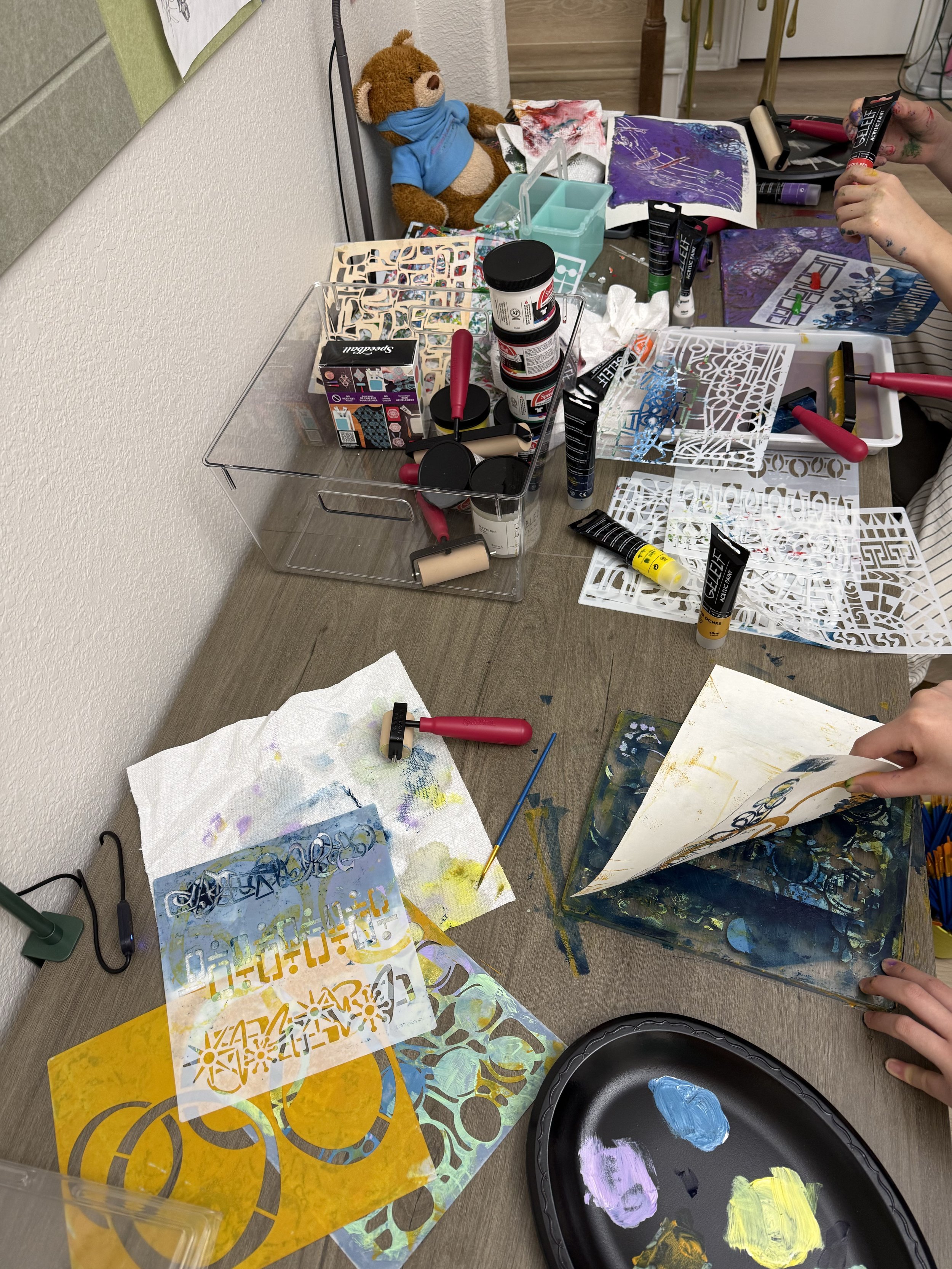 A cluttered art and craft workspace with paints, stencils, brushes, paper, and a stuffed bear wearing a face mask. A person's hands are seen handling a paper or stencil, and another hand is holding a tube of paint.