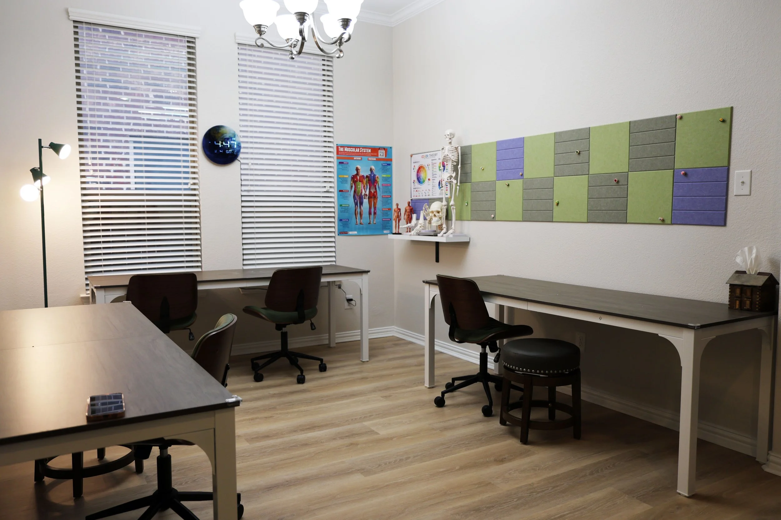 A small medical consultation or therapy room with three desks, four chairs, a tissue box, and educational posters on anatomy and the nervous system. The room has three windows with blinds, a round wall clock, a floor lamp, and a decorative wall with 