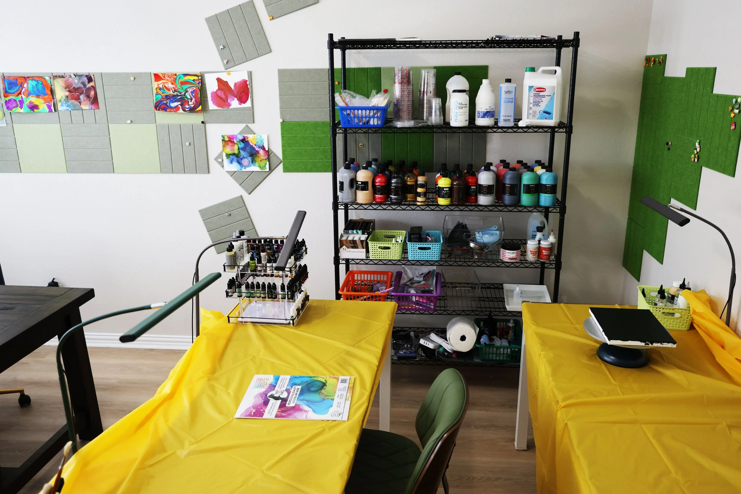 Indoor art studio with tables covered in yellow cloth, tools, and supplies, metal shelving with paints and containers, colorful artwork on the walls, and green decorative wall elements.