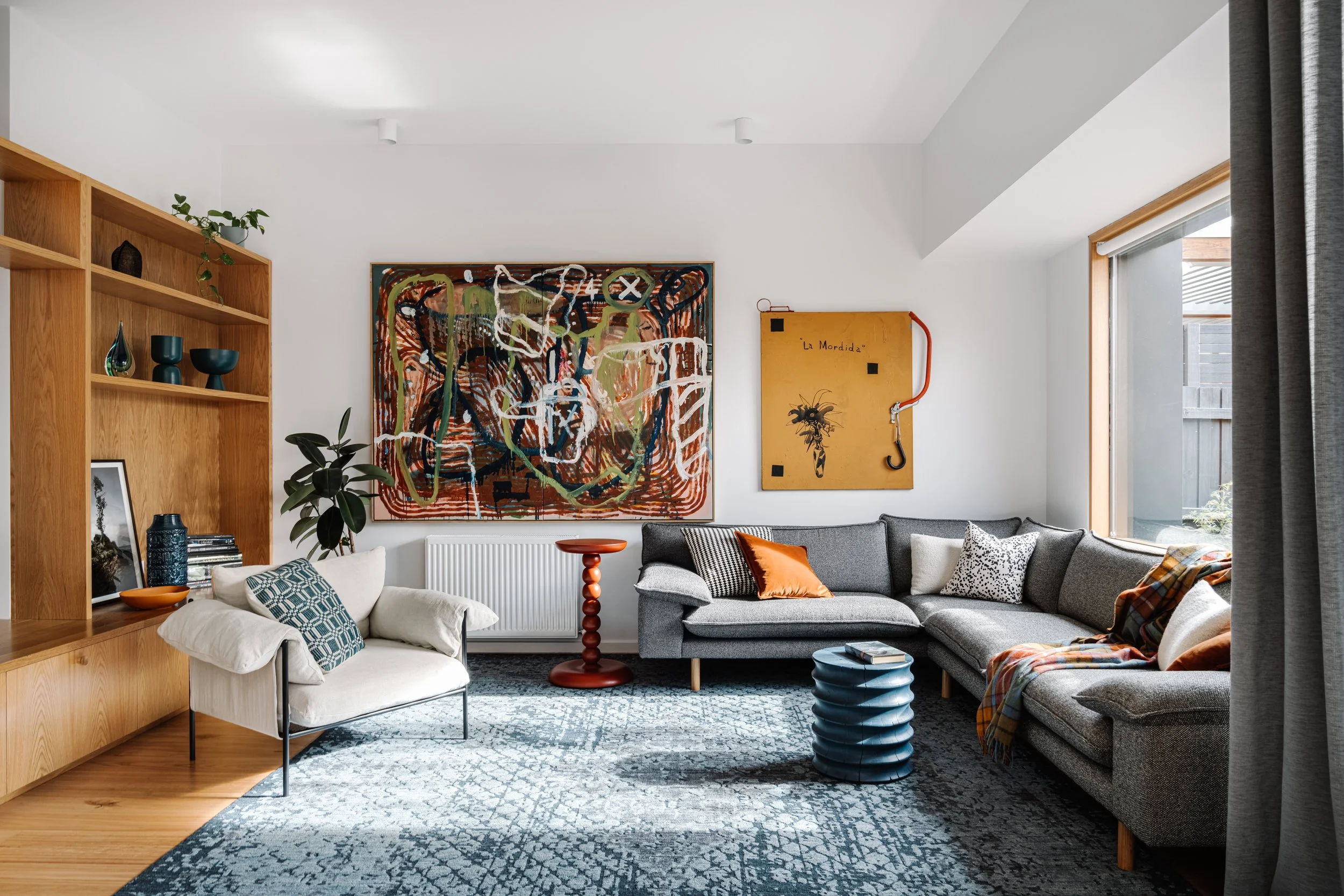 Living room with a large abstract painting on the wall, a gray sectional sofa with pillows, a white armchair, a red side table, a tall bookshelf with decorative items, a window with curtains, and a blue rug.