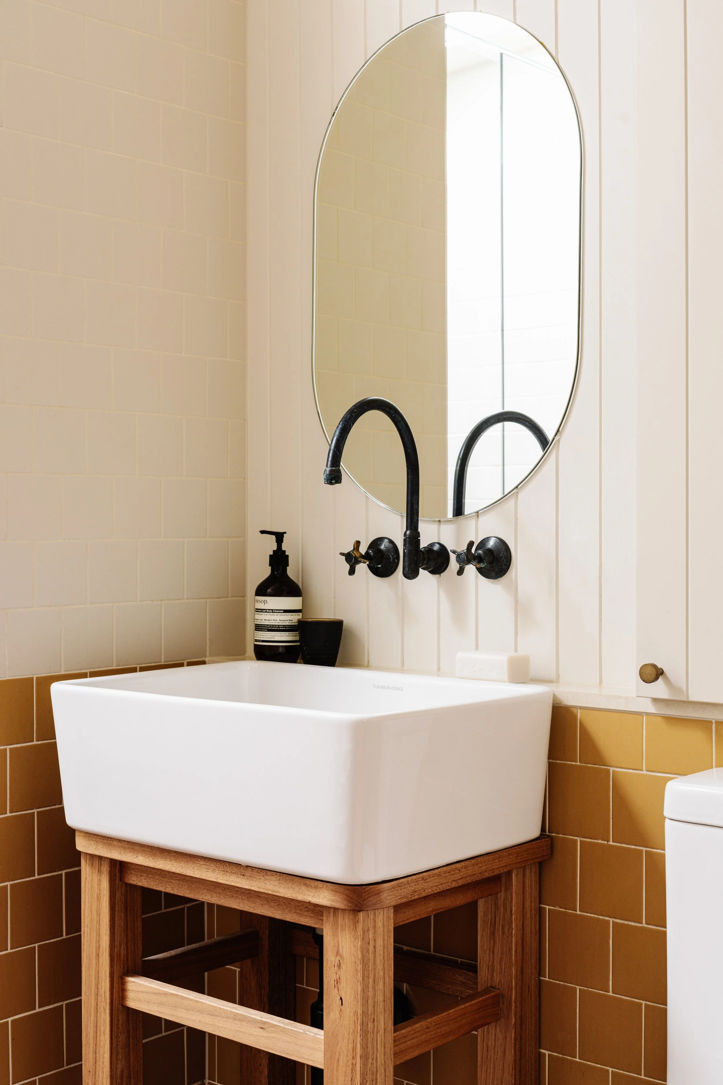 A white rectangular sink with a wooden stand underneath, set against a tiled wall, with a mirror above it reflecting a window and part of the bathroom