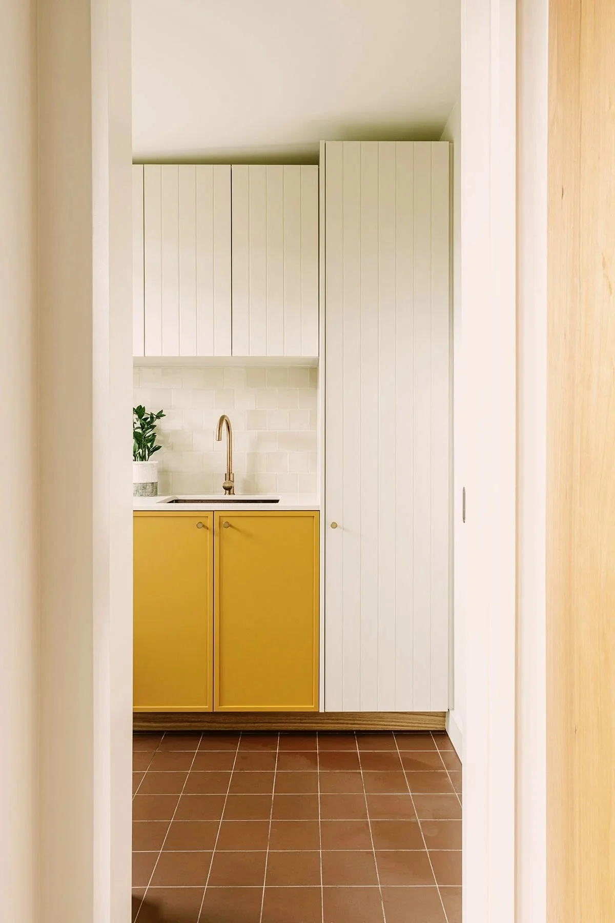 Small kitchen with yellow lower cabinets, white upper cabinets, a white backsplash, and a pinkish gold faucet. There is a potted plant on the counter.