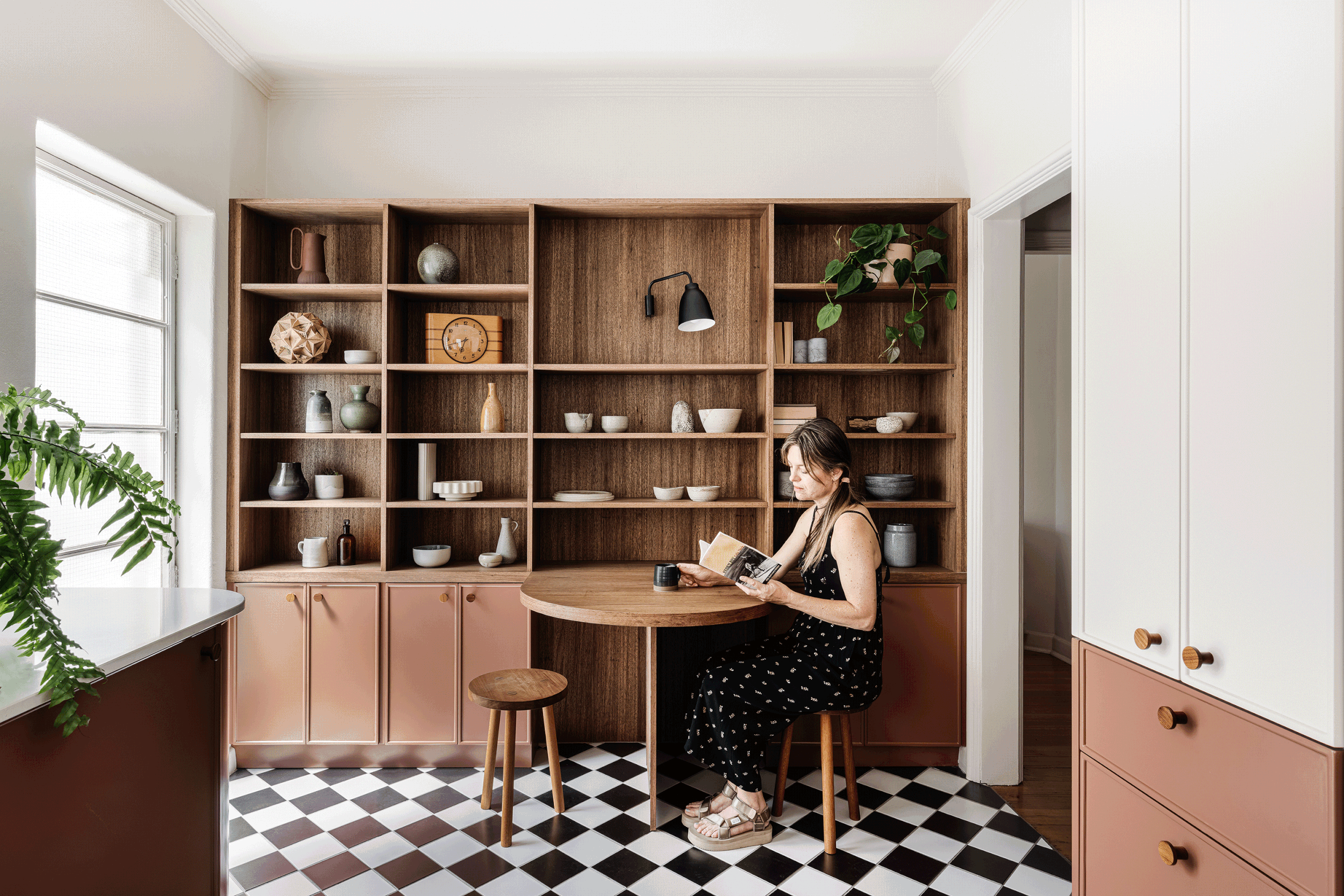 Woman sitting on a wooden stool in a modern kitchen with wooden shelves, white and pink cabinets, and black and white checkered floor, reading a magazine.