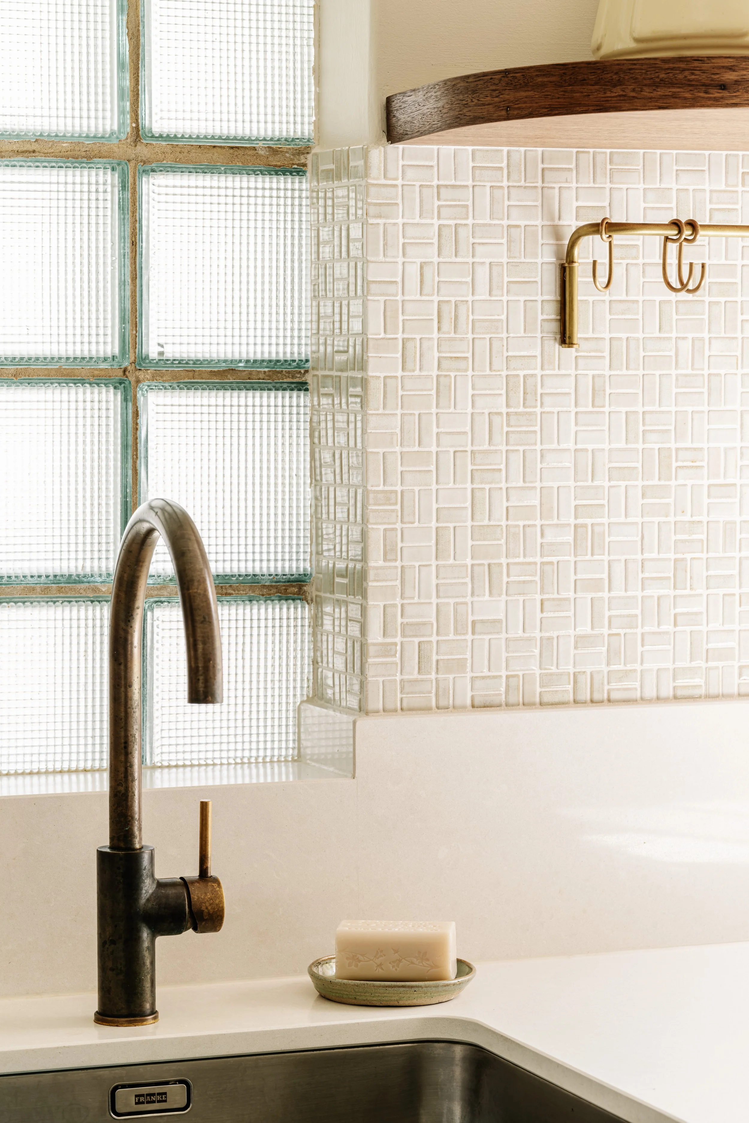 A kitchen sink with a dark metal faucet, a bar of soap in a green dish, glass blocks window, tiled wall, and a small soap holder.