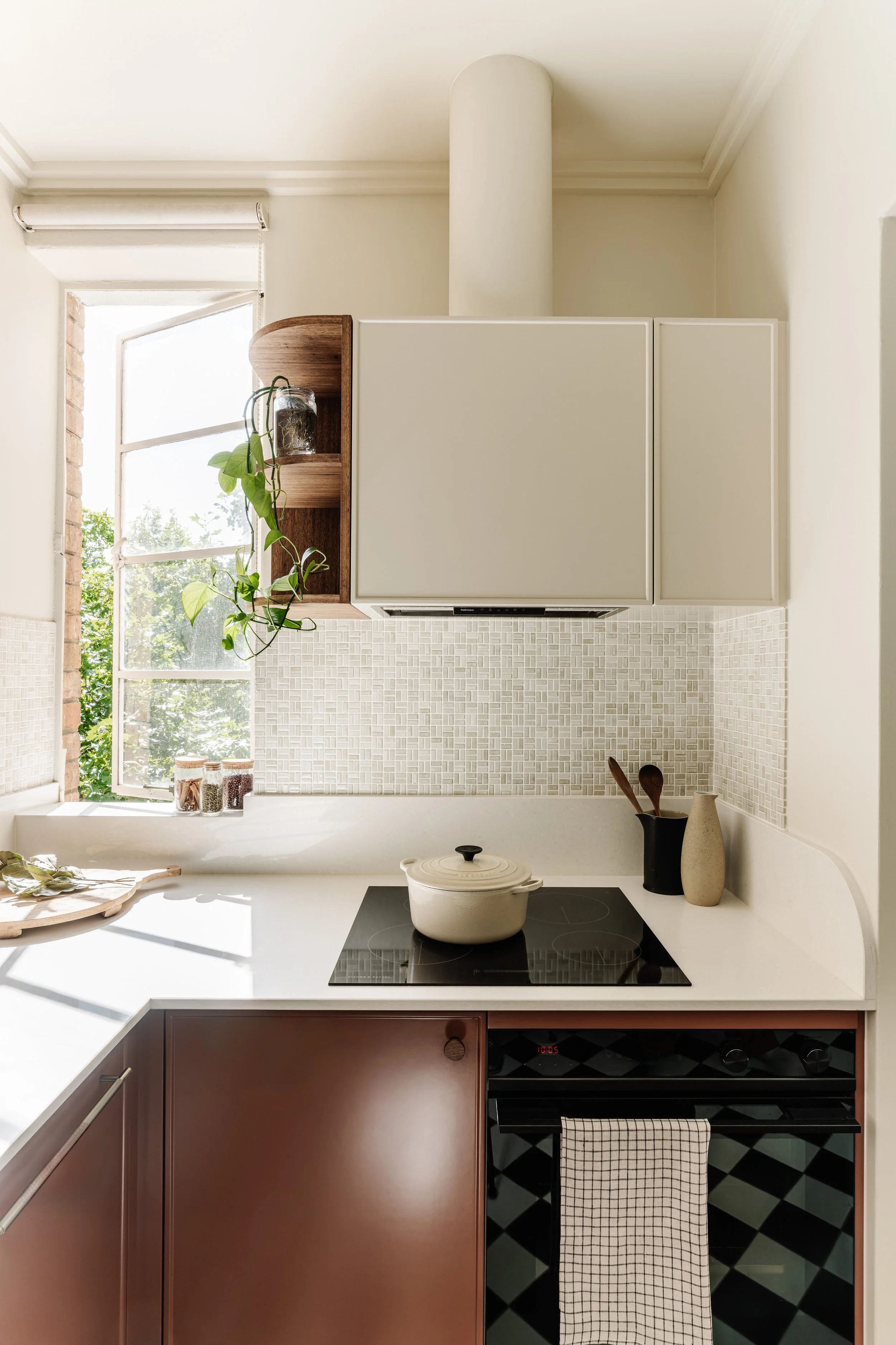 A modern kitchen with a window, white countertop, countertop stove with a pot, brown and black cabinets, a black oven, a cloth towel hanging, and decorative items including vases and plants.