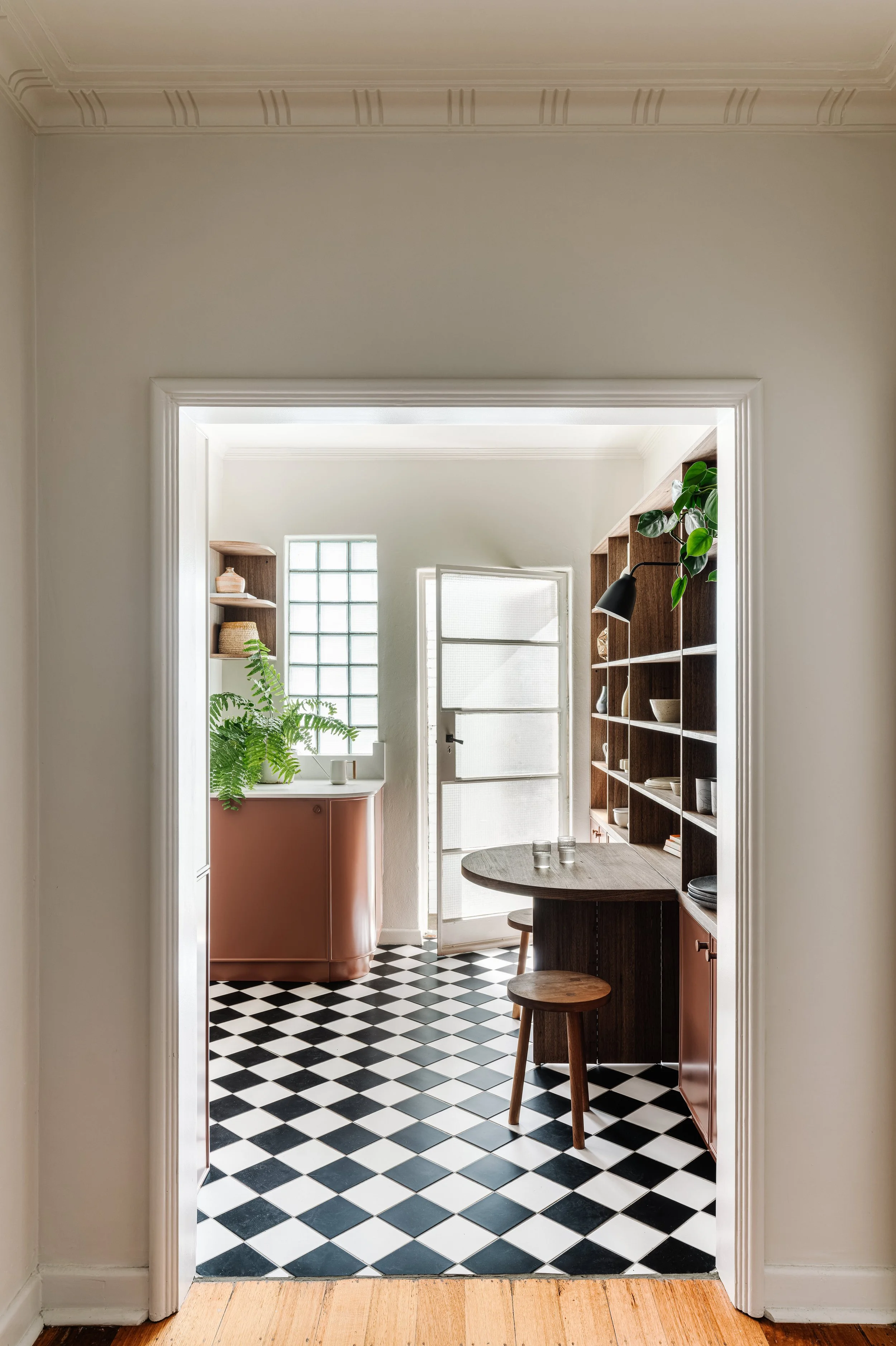 View through doorway of a kitchen with black and white checkered floor, a pink cabinet, wooden shelves with decorative items, and a wooden table with stools.
