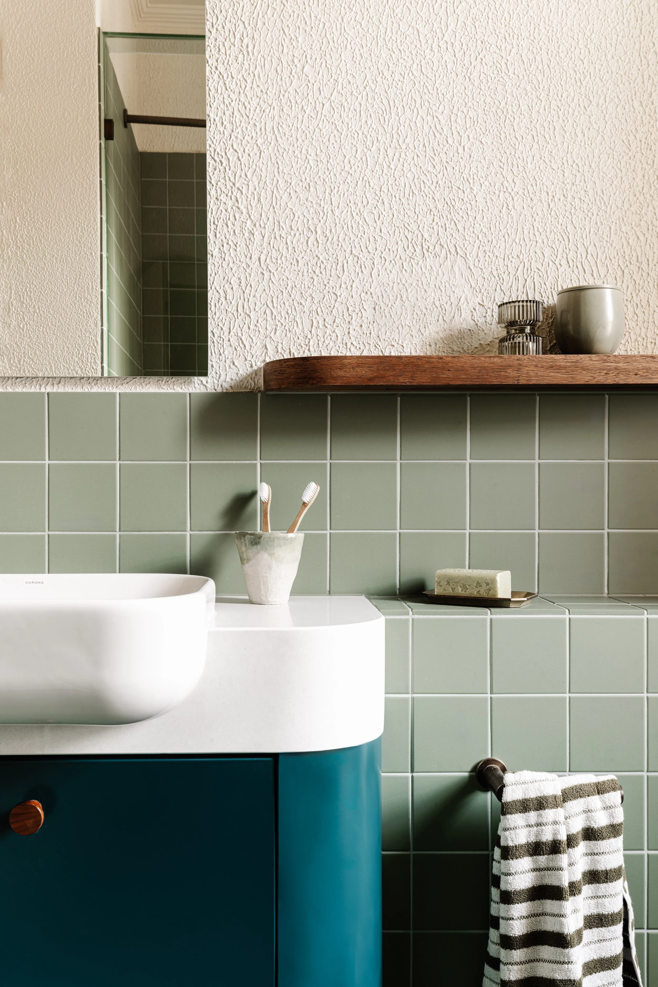 Bathroom with green tiled wall, white textured wall, wooden shelf with decorative items, toothbrushes in a holder, towel hanging on a rod, and part of a mirror.
