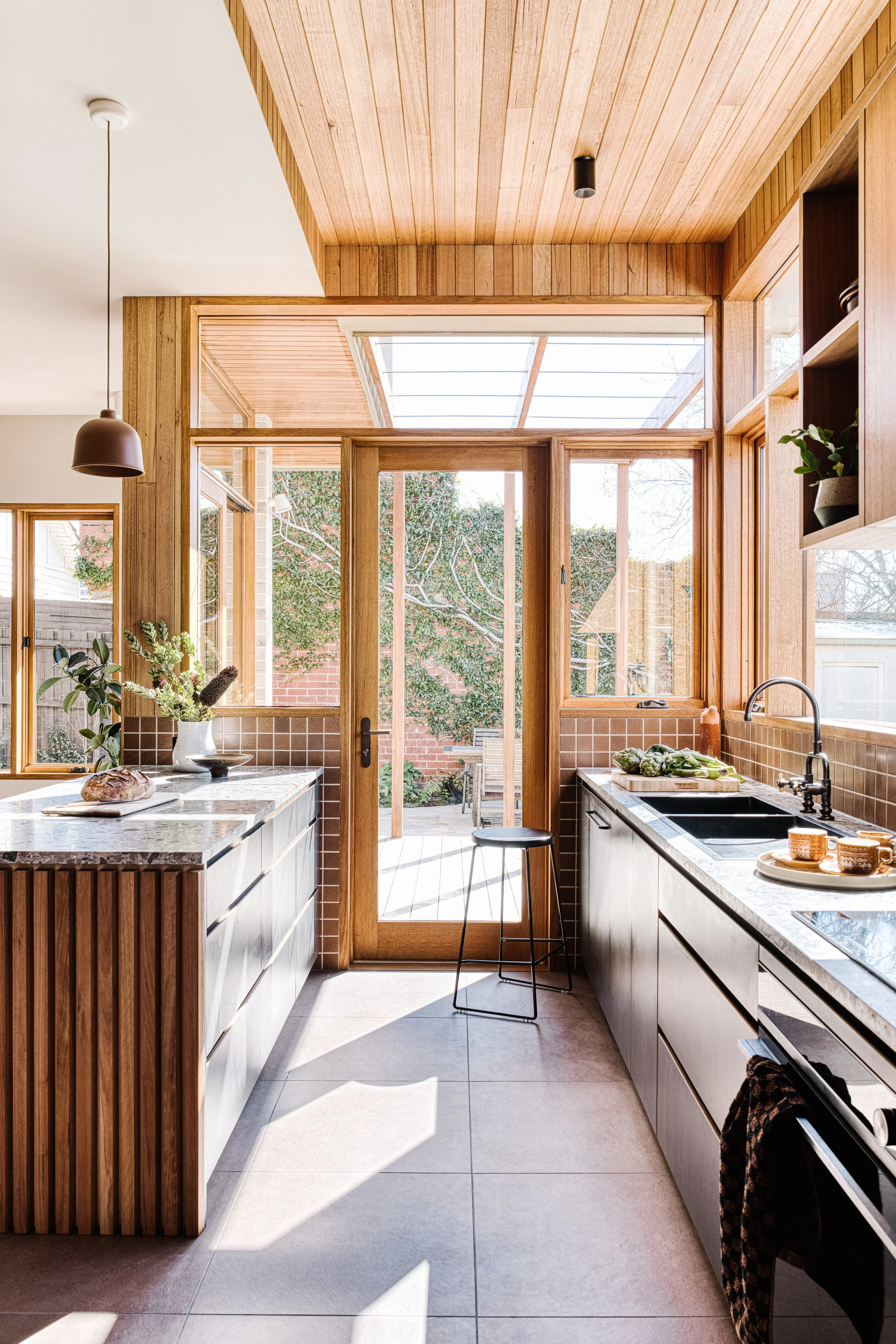 A modern kitchen with wooden cabinets, large windows, and a granite countertop. Outside view includes trees and a brick wall. There are plants and kitchenware on the counter, with sunlight streaming in.