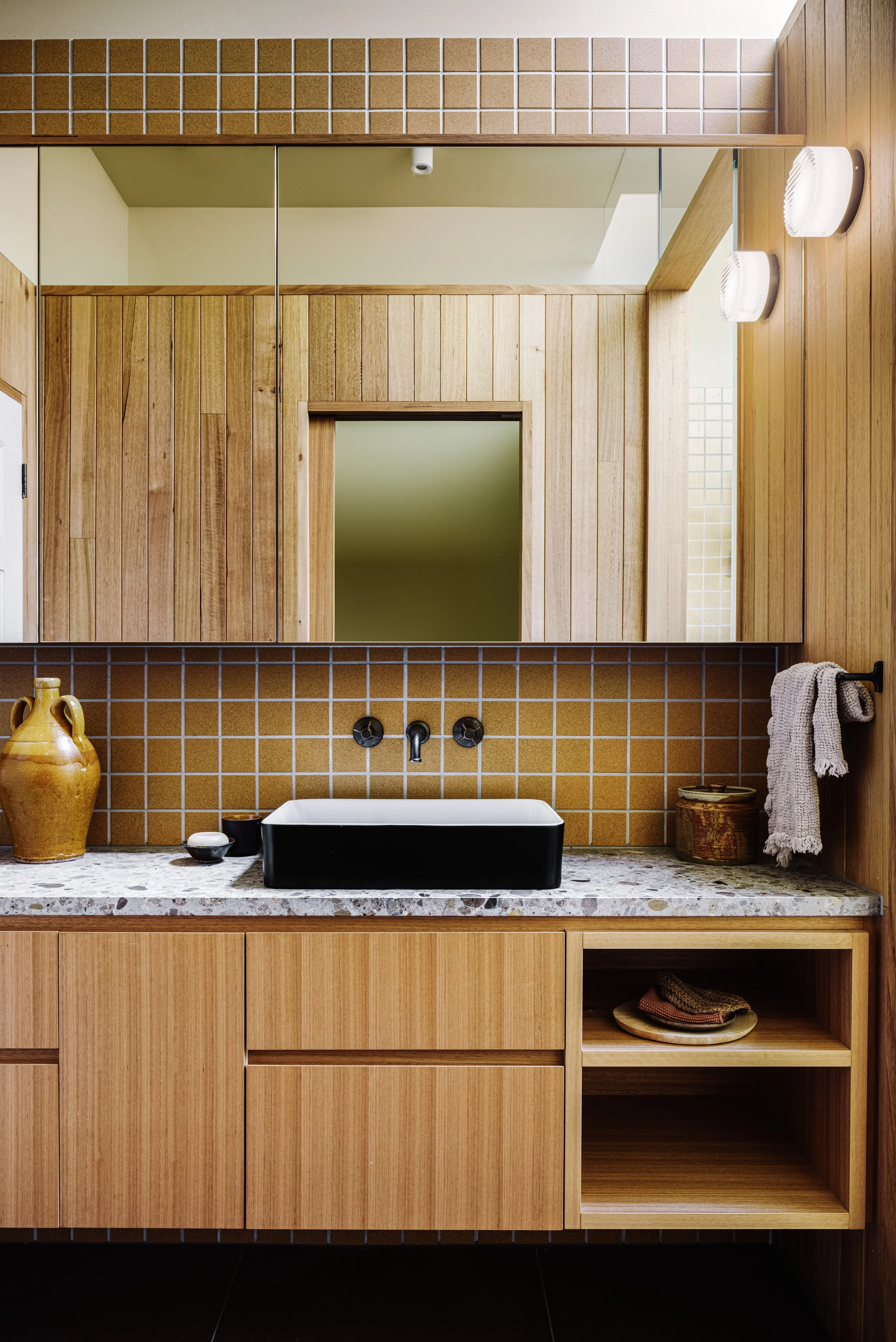 A modern bathroom vanity with a black rectangular vessel sink mounted on a speckled granite countertop, wooden cabinets below, a large mirror above, and warm wood and yellow tile accents.