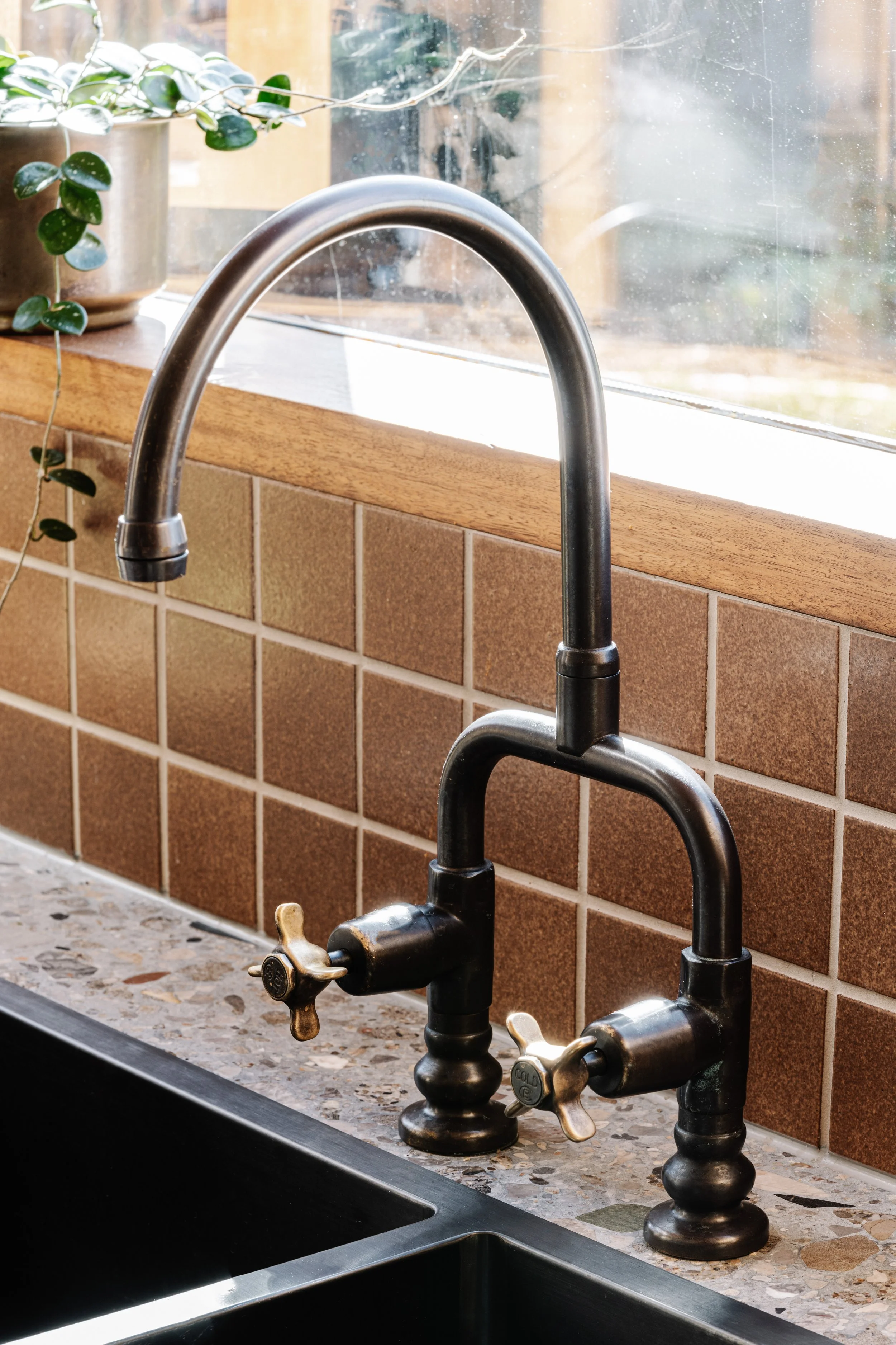 Close-up of a kitchen sink faucet with a window, potted plant, and tiled backsplash in the background.