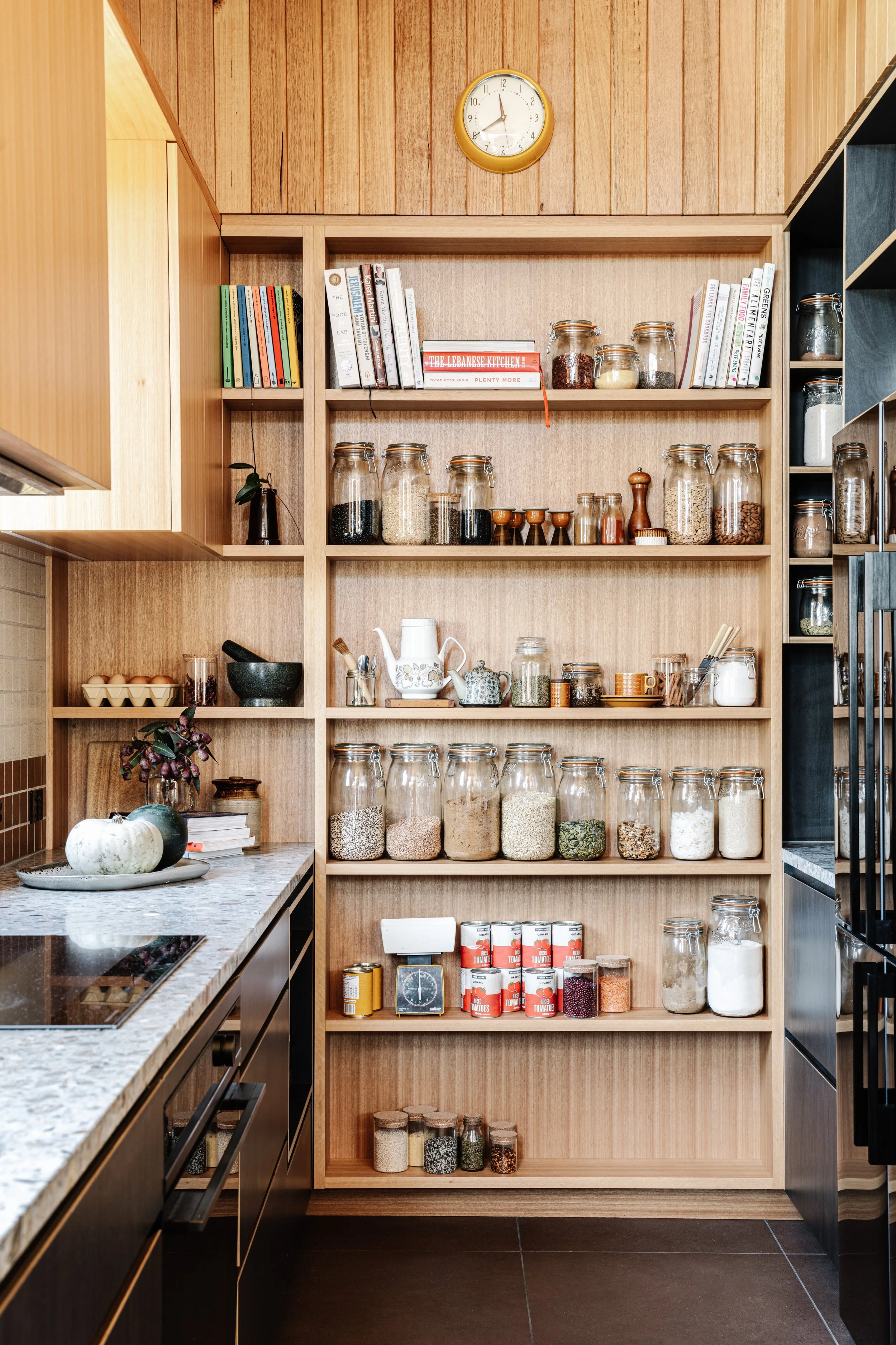 A kitchen shelf filled with jars of spices, grains, and dried foods, with cookbooks and a clock above, and a countertop with eggs and decorative pumpkins in front.