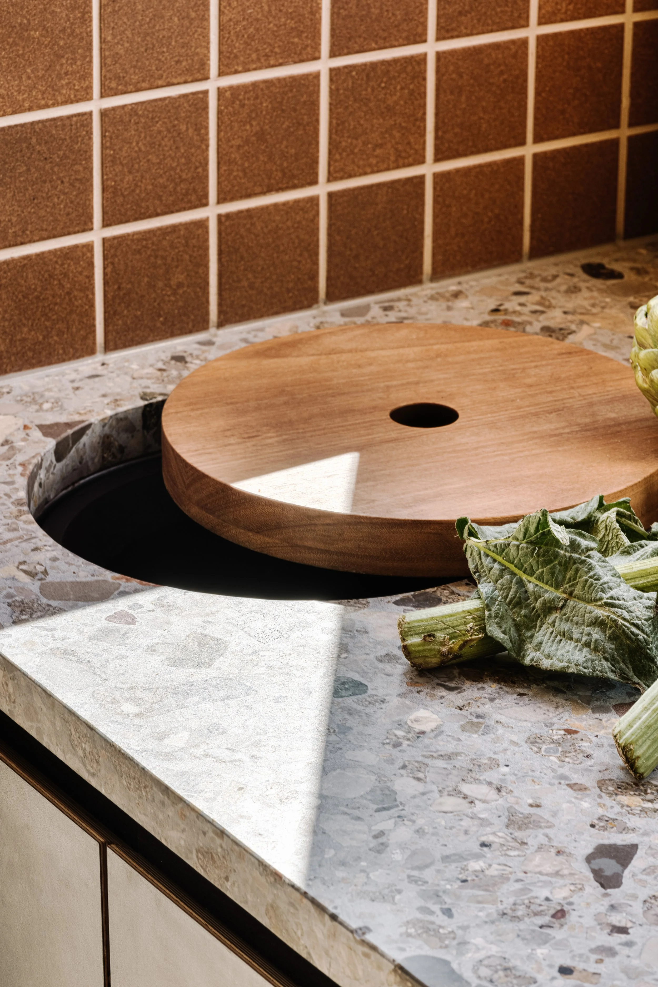 A wooden serving tray with a hole in the center, placed on a speckled stone countertop, with wilted greens and artichokes nearby, against a background of brown tiled wall.