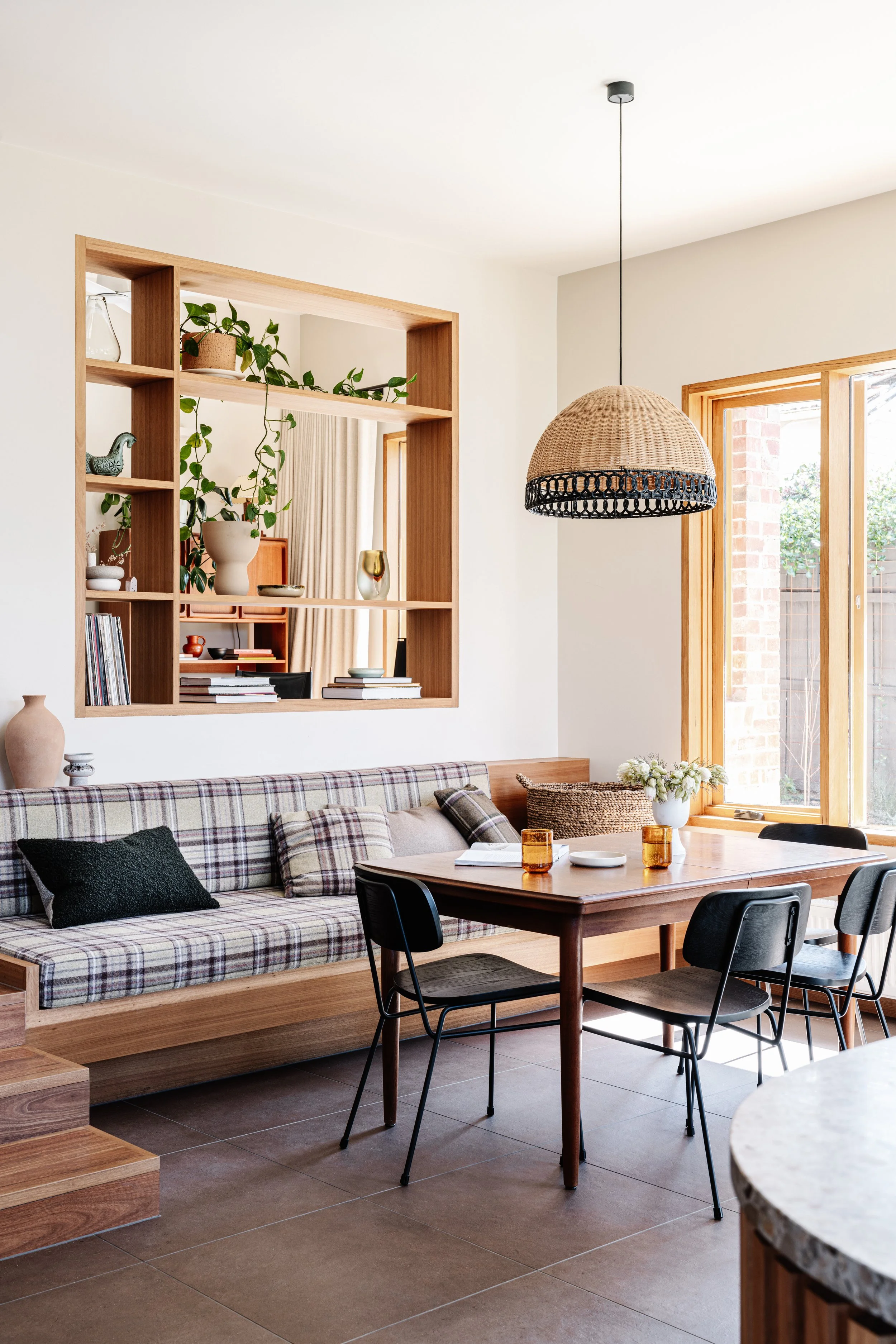 A cozy, modern dining area with a wooden table, black chairs, and a plaid cushioned bench. There is a large window with natural light and a decorative hanging rattan lamp.