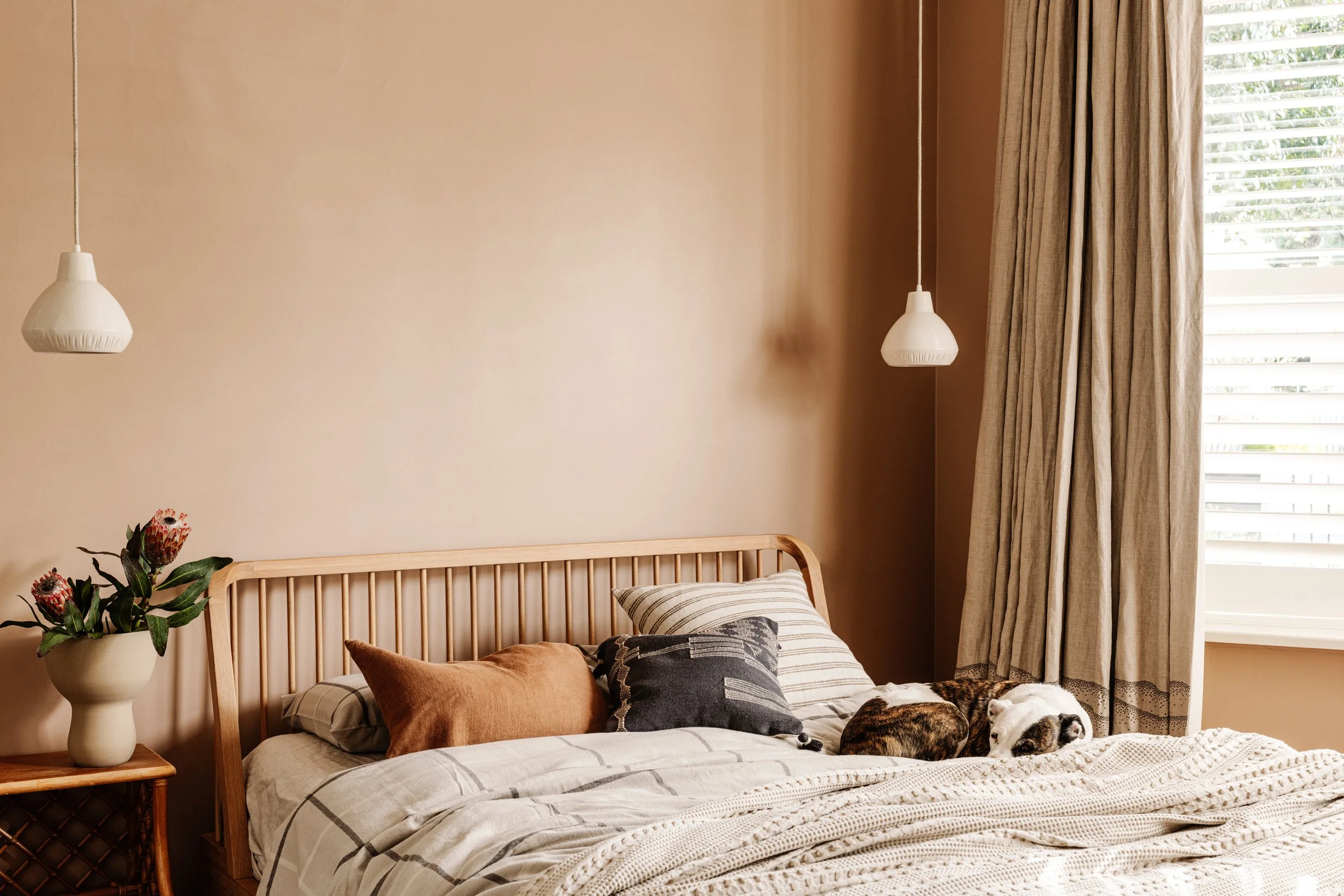 A cozy bedroom with a wooden bed frame, striped bedding, and various pillows. A vase of flowers sits on a side table, and there are two hanging pendant lights above. The room has beige walls, beige curtains, and a window with blinds letting in natural light.