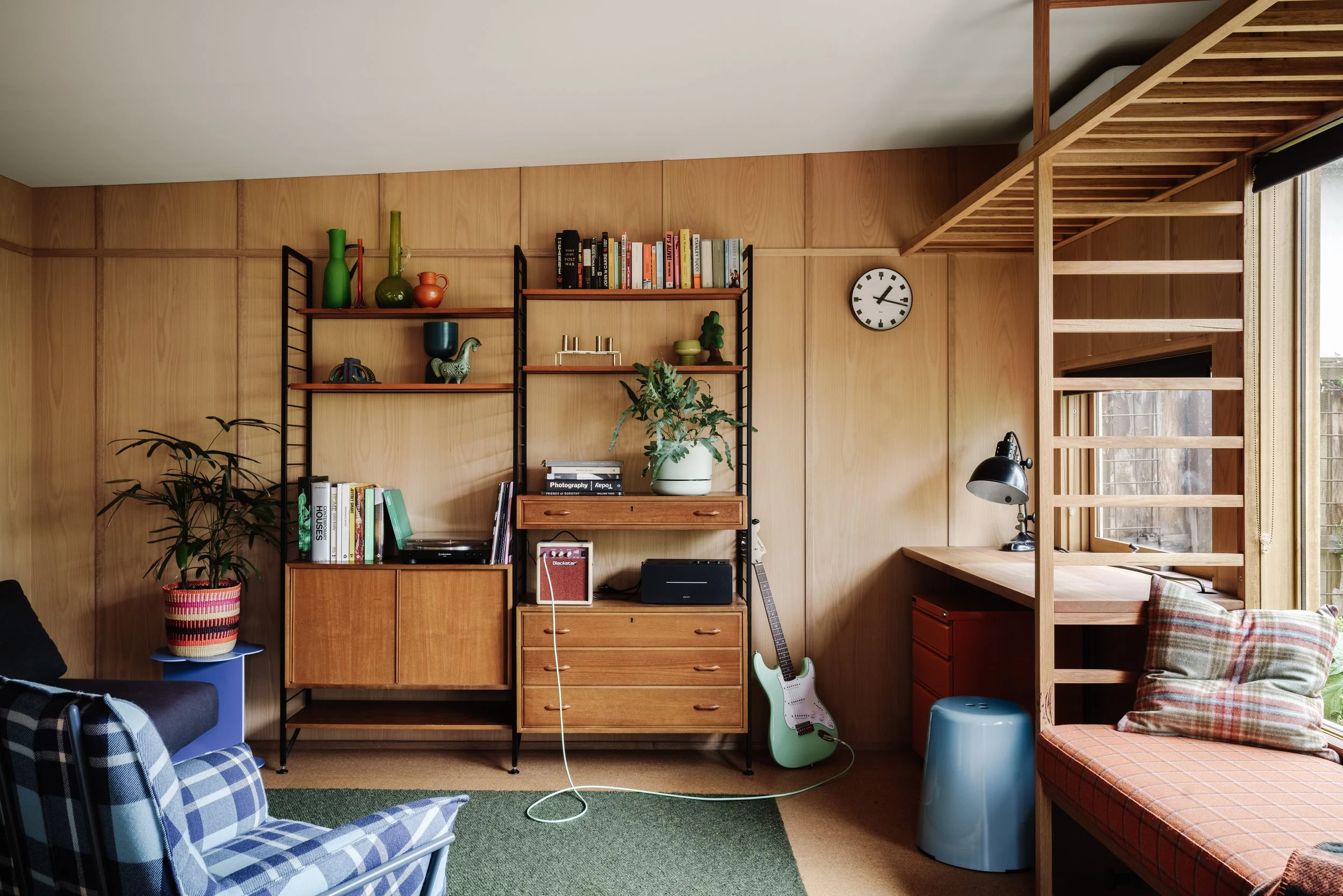 Interior of a cozy living space with wooden panel walls, a bookshelf with books, decorative vases, a plant, a guitar, and a window with natural light.