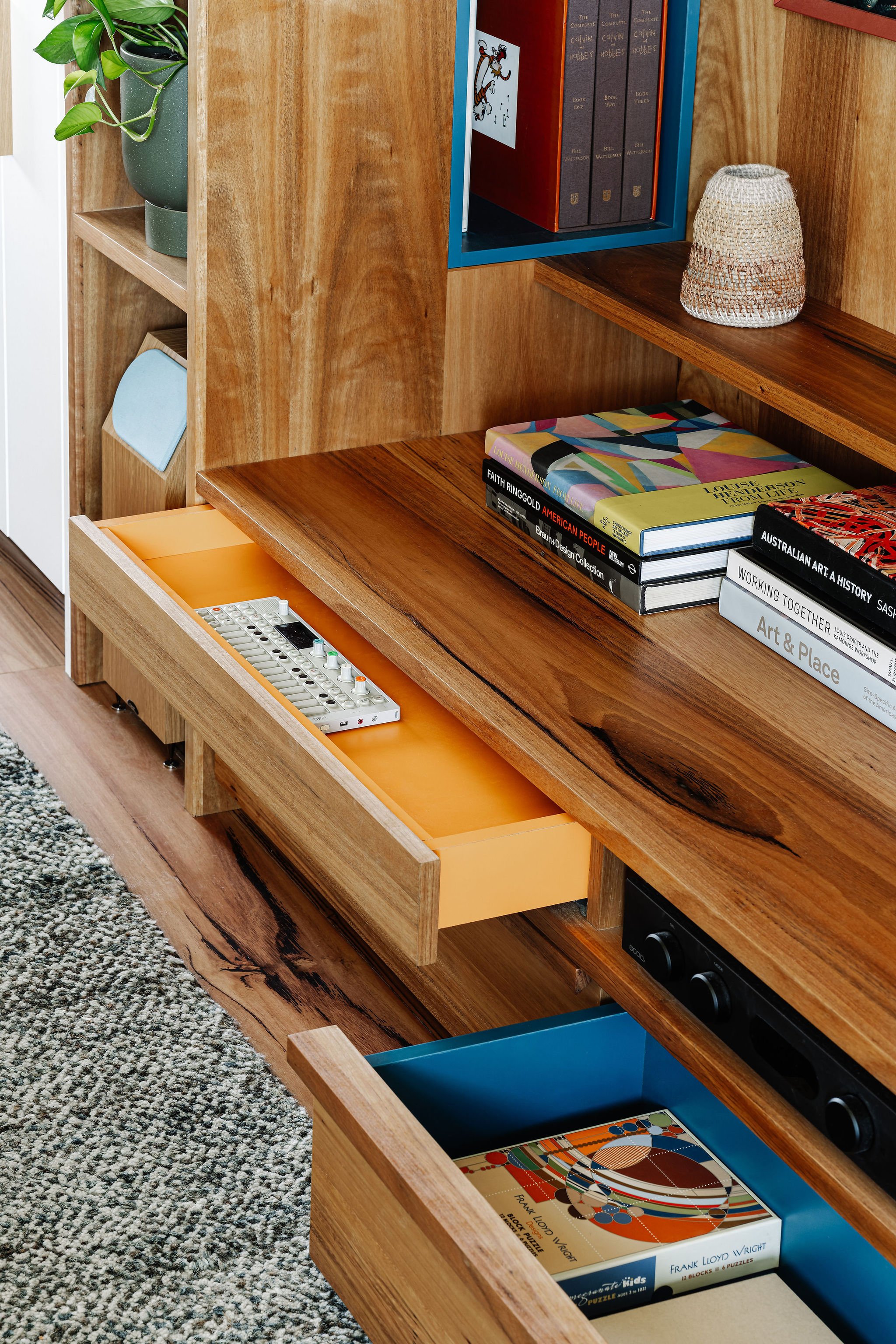 Open wooden drawers and shelves containing books, a plant, a ceramic vase, and a puzzle box, with a patterned carpet nearby.