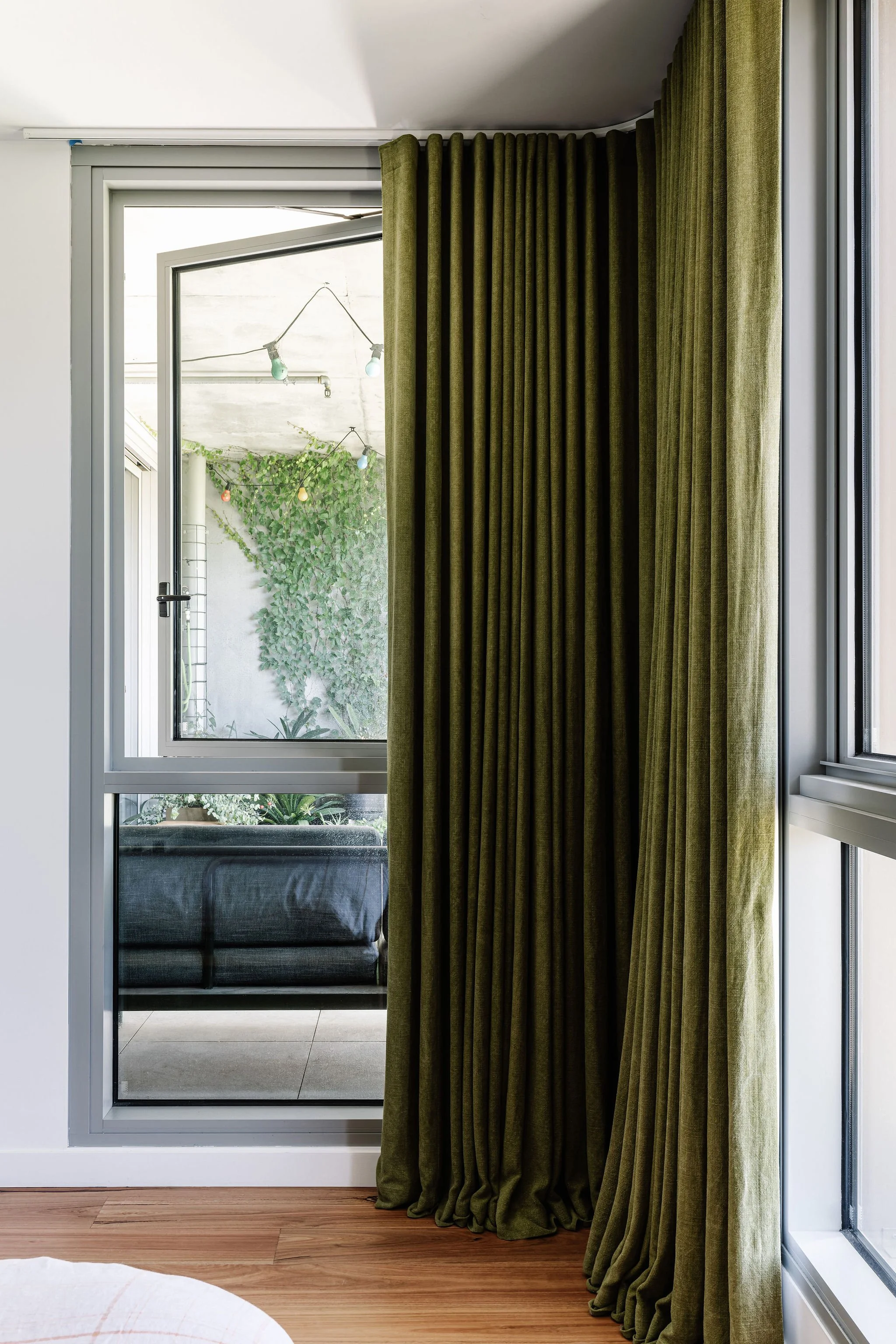 A rectangular glass door with a sliding screen, partially covered by a green velvet curtain, leading to a balcony with outdoor plants and string lights.