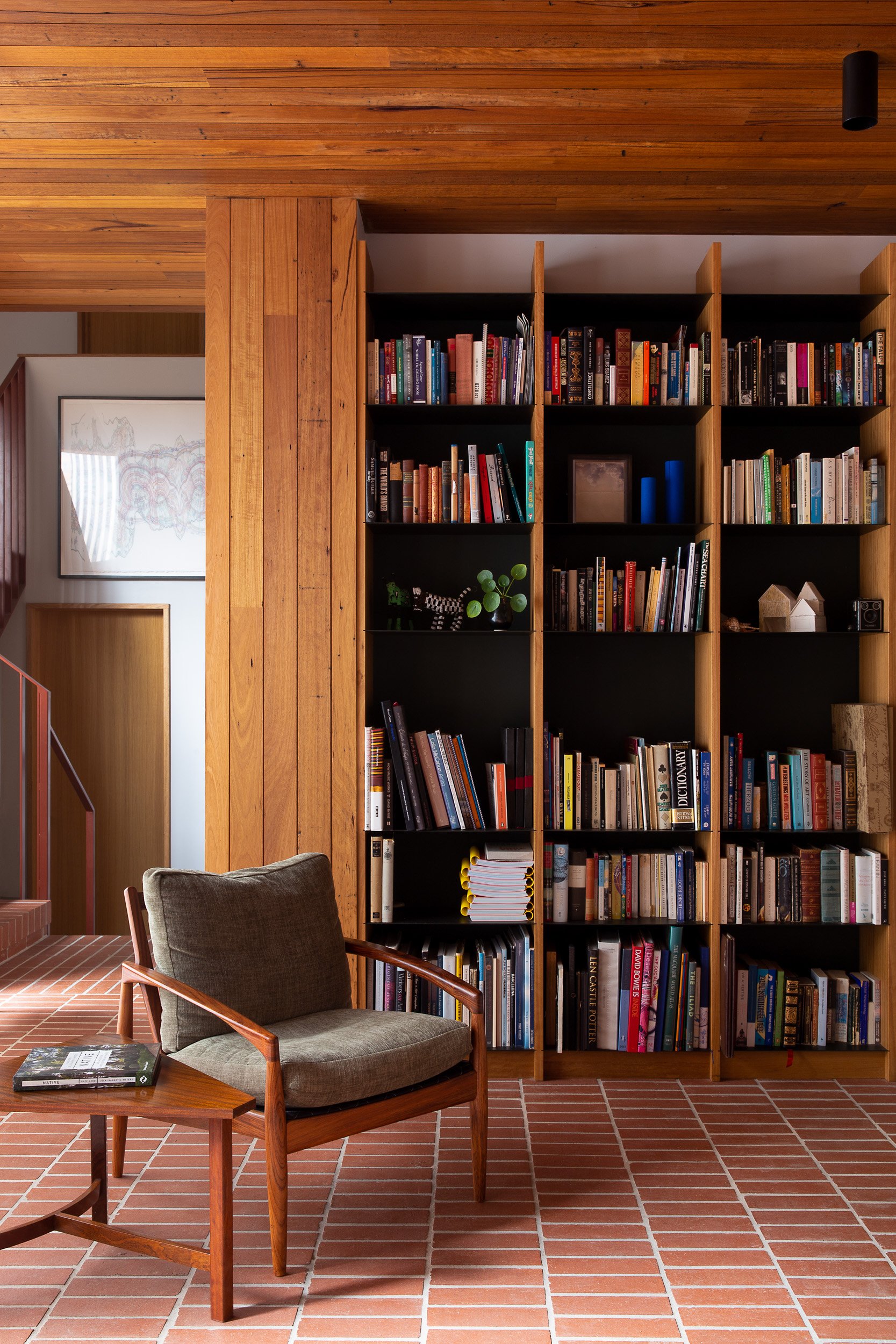 Living room featuring a large wooden bookshelf filled with books, a wooden armchair with a cushion, a small side table with a book on it, and a brick floor. The ceiling and a wall are also made of wood.