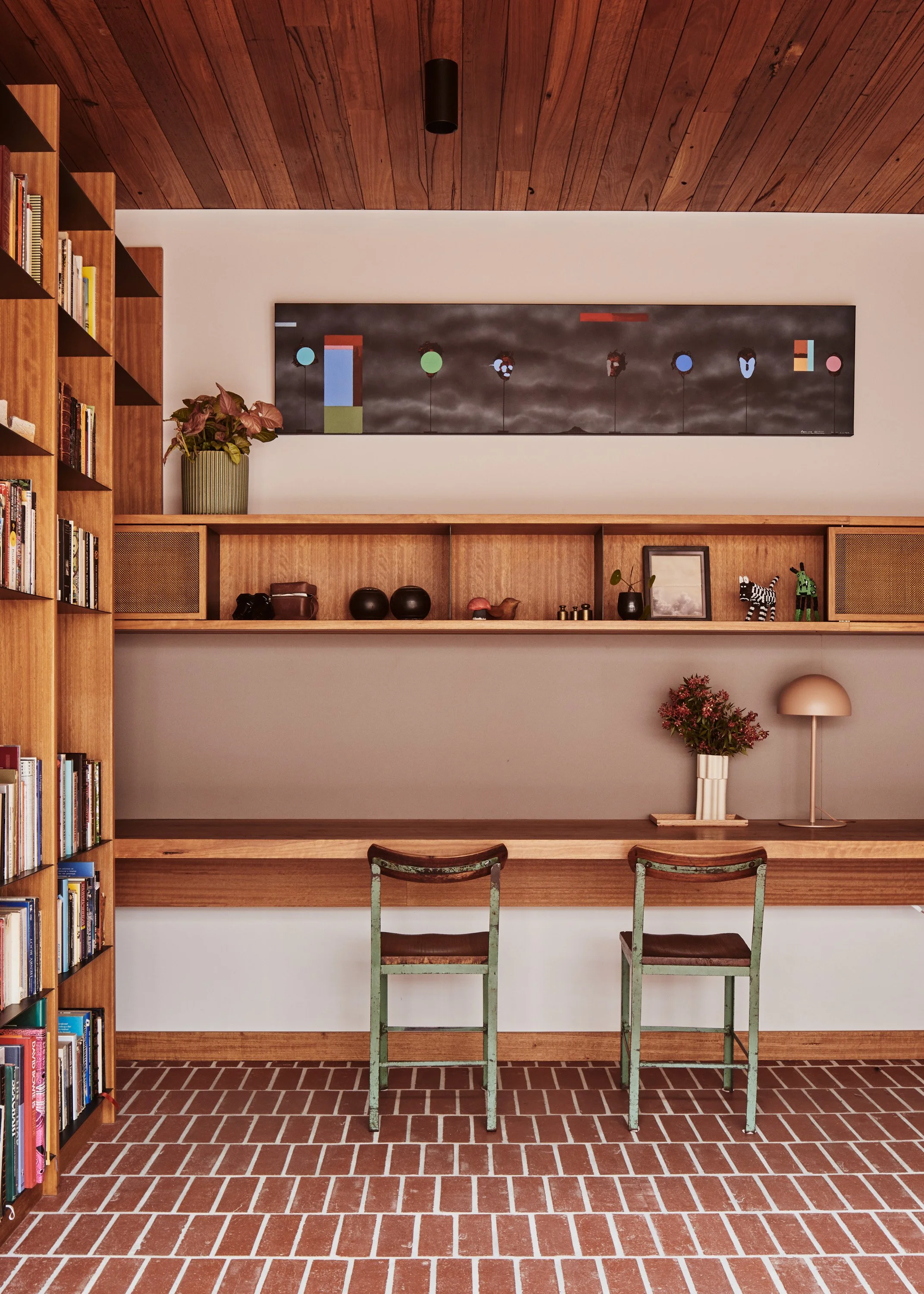 Living room with wooden ceiling, built-in wooden bookshelf on the left filled with books, a long wooden shelf with various decorative items including a potted plant, picture frame, and small sculptures. Two worn, green wooden chairs sit in front of a white wall with a floating wooden shelf, a lamp, and a vase with flowers. Brick flooring is visible in the foreground.