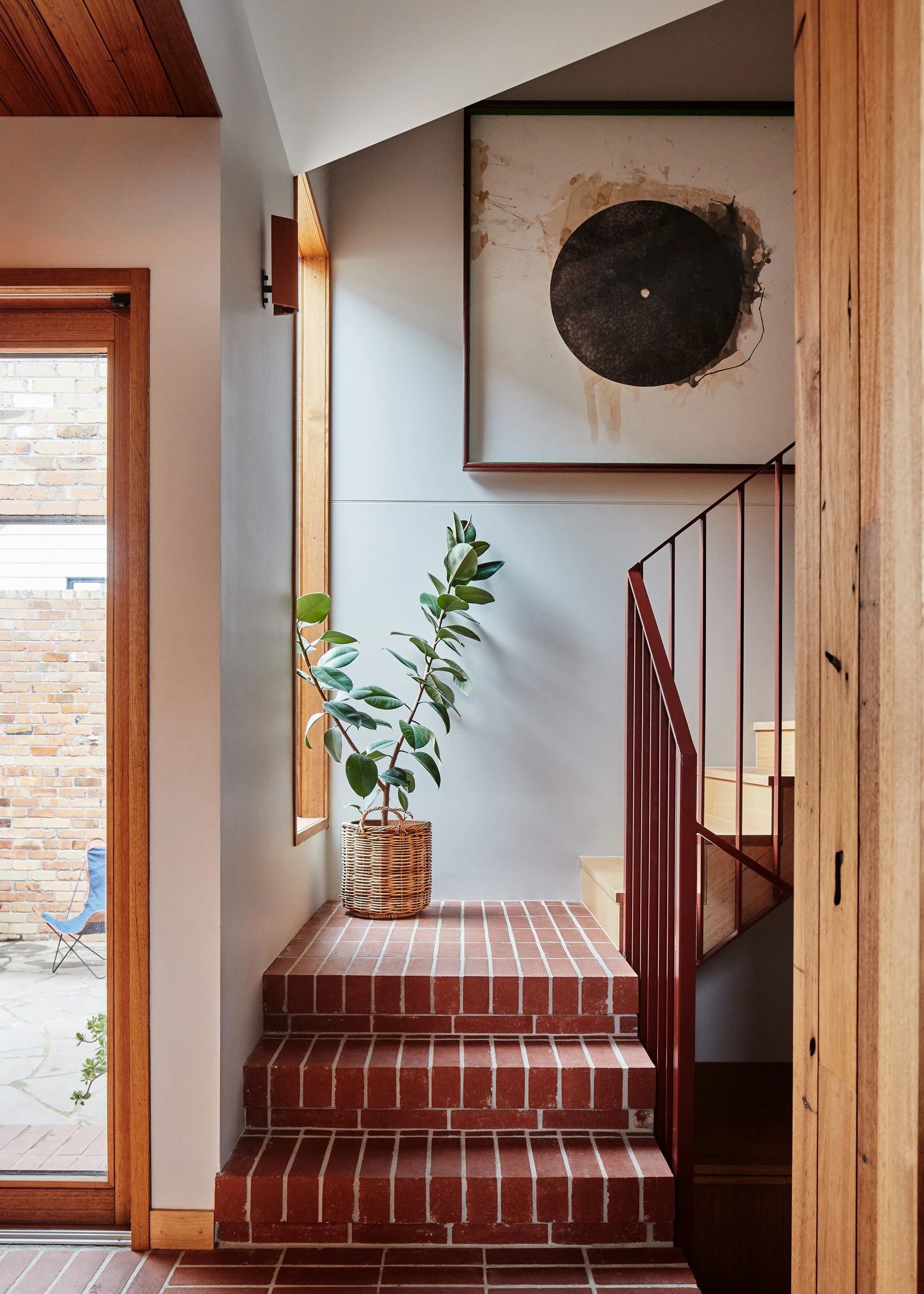 Interior of a house with brick stairs, a potted plant, a framed artwork, and a wooden staircase with a red railing.