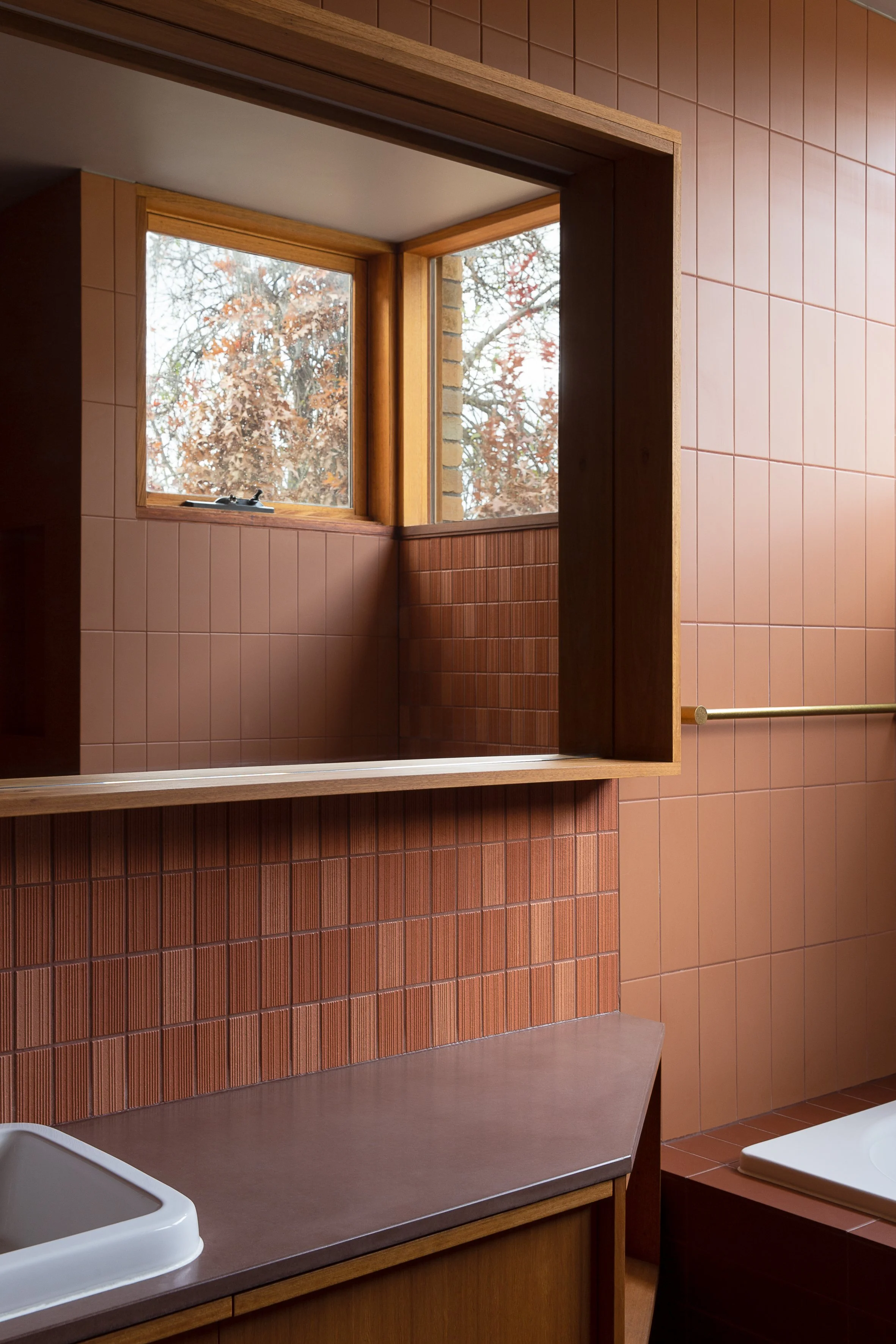 A view of a bathroom with pink tiled walls, a large window showing trees outside, a ceramic sink, and a brown countertop.