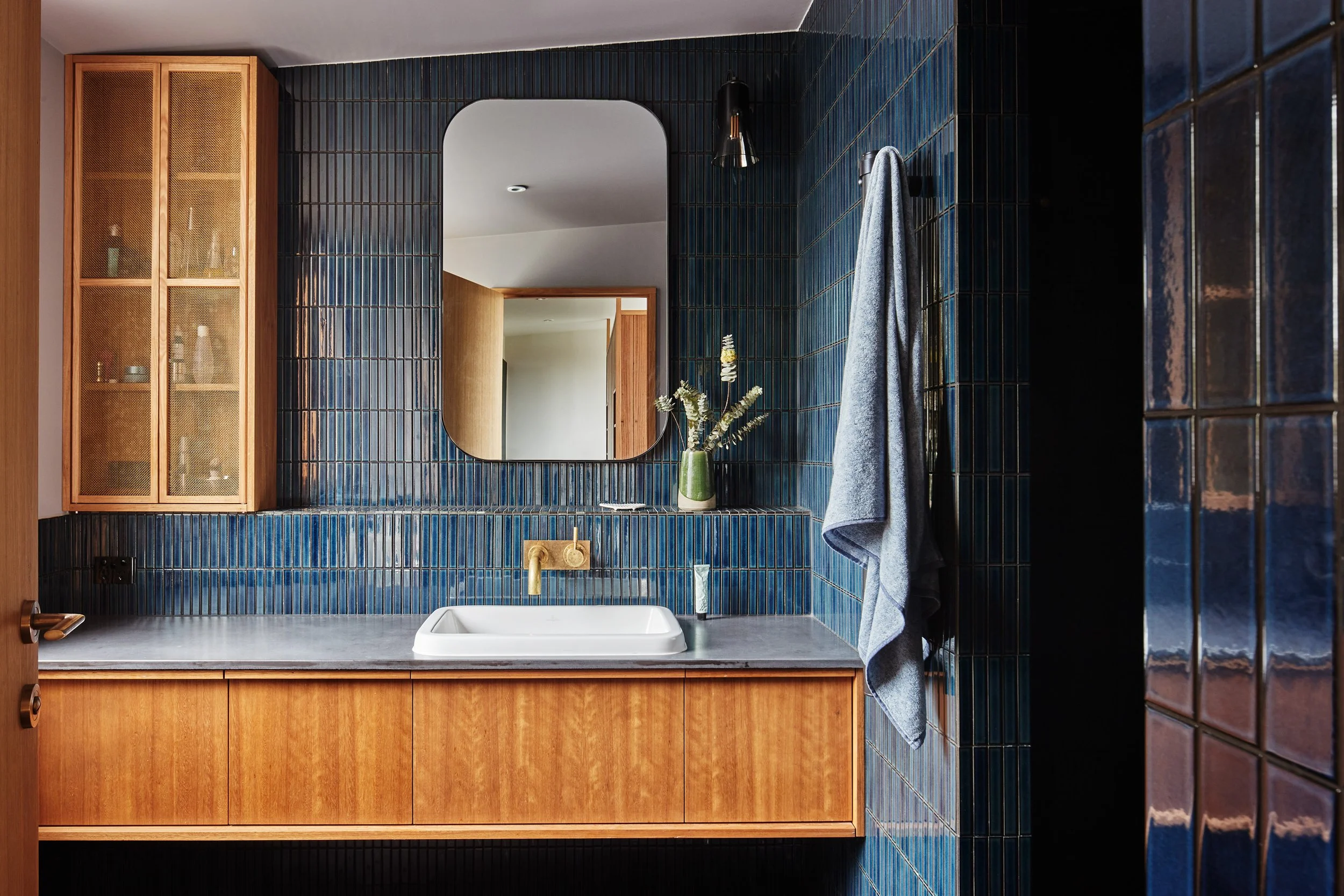 A modern bathroom with blue tiled walls, a rectangular white sink, a gold faucet, a wooden cabinet below the sink, a mirror on the wall, a decorative vase with flowers, a gray towel hanging on a hook, and a black wall sconce light.
