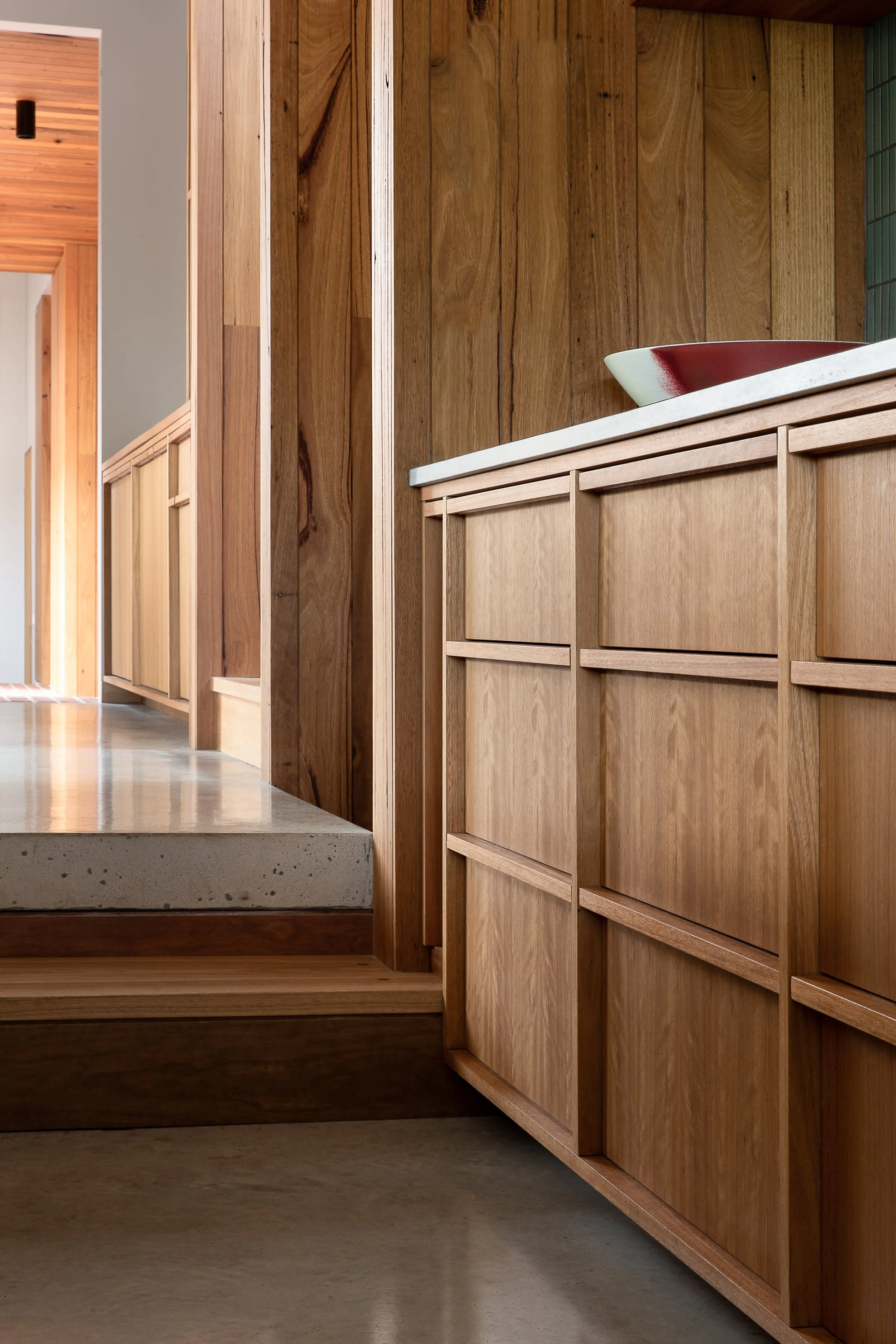 Interior view of a modern home with wooden cabinetry and a white countertop. A decorative bowl with red and white colors sits on the countertop. The space features warm wooden accents and a polished concrete floor.