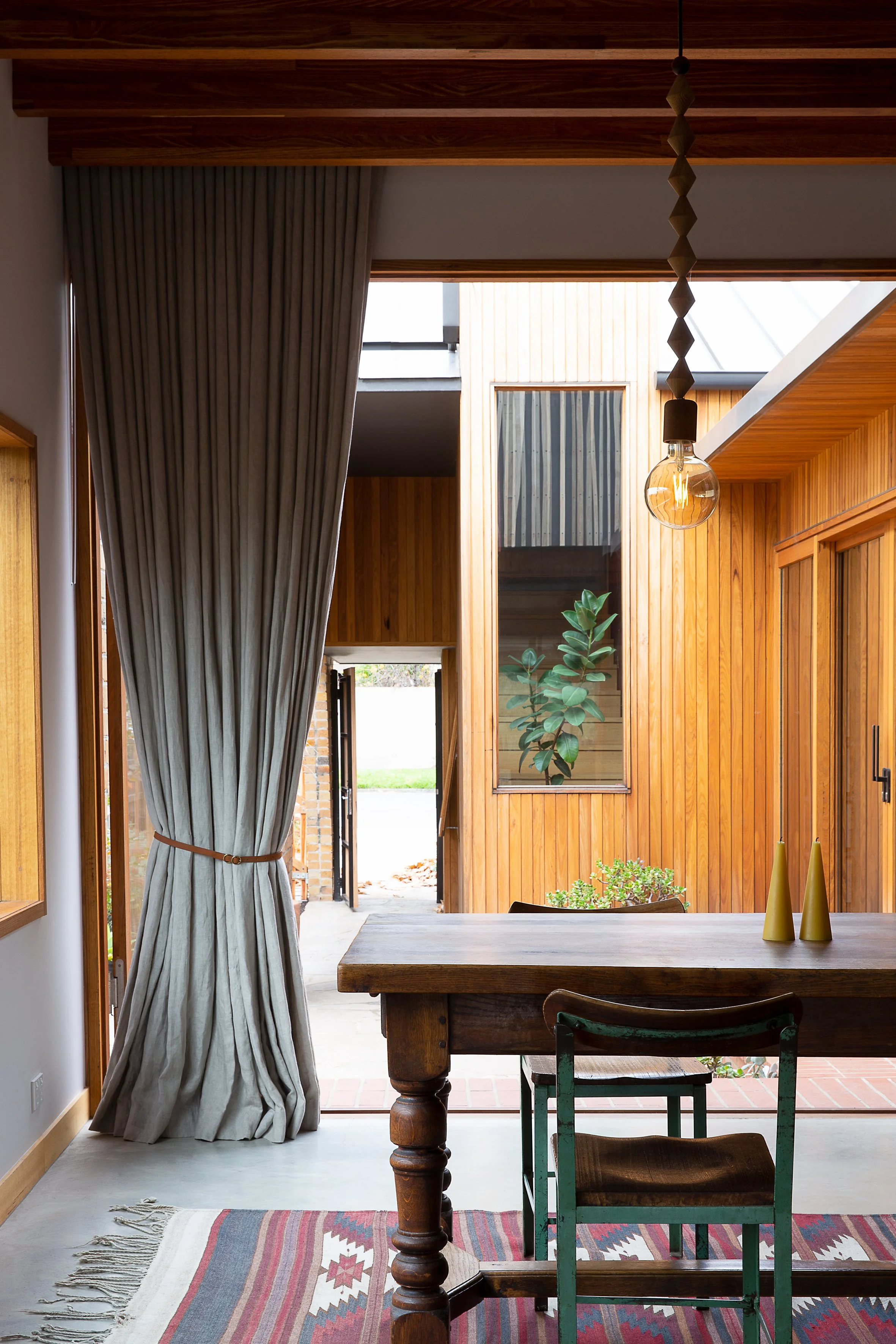 A dining area with a wooden table, two chairs, a colorful rug, beige curtains, and an open door leading outside, with wooden panel walls and a large window.